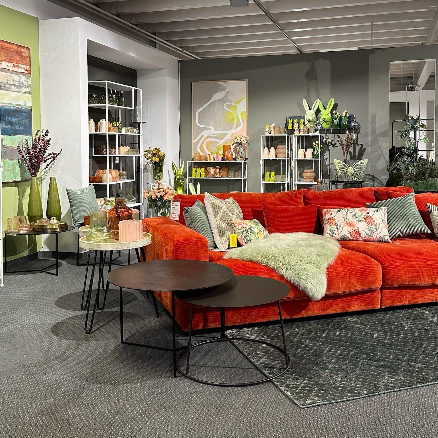 Moderne Wohnzimmer-Ausstellung mit rotem Cord-Sofa, schwarzen Beistelltischen und frischer Frühjahrsdeko in Grüntönen.