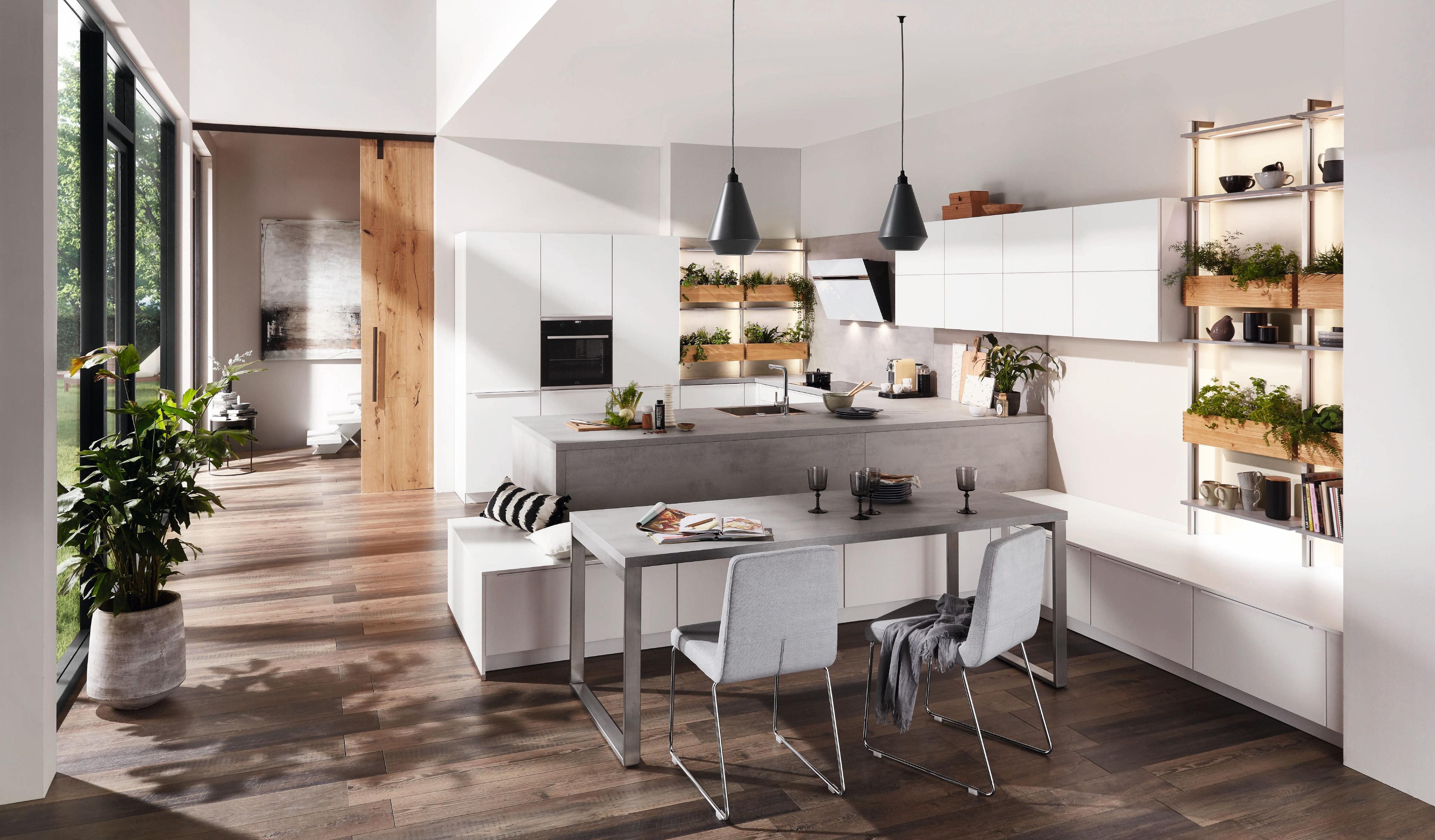Moderne Küche mit weißen Hochglanzfronten, einer Betonarbeitsplatte, offenen Holzregalen mit Pflanzen und einem stilvollen Essbereich mit minimalistischen Möbeln und Holzboden.