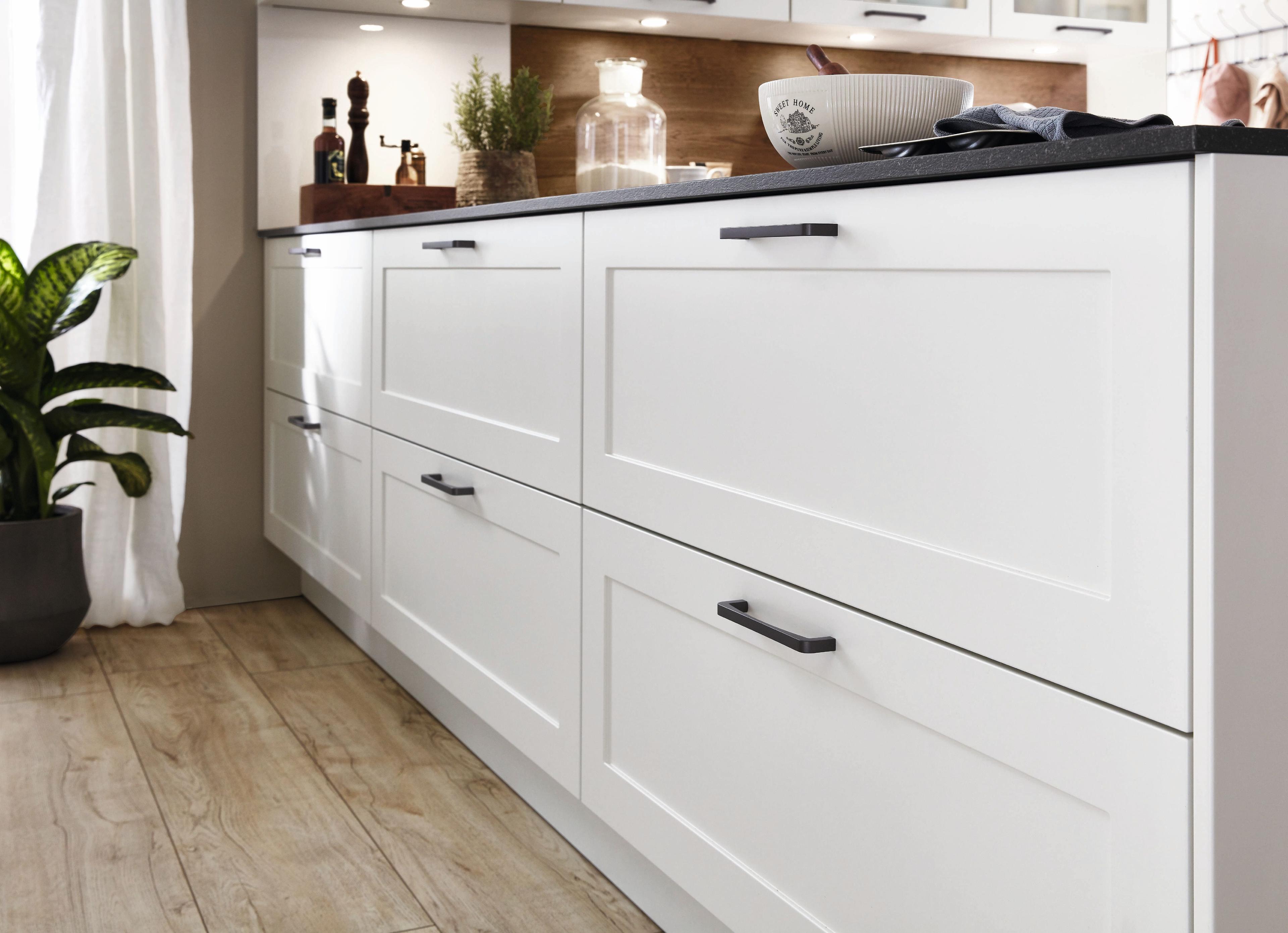 Detailaufnahme einer modernen weißen Küchenzeile mit eleganten, schwarzen Griffen und Holzregalen. Auf der Arbeitsfläche sind Gewürzmühlen, eine Pflanze und eine Schüssel sichtbar.