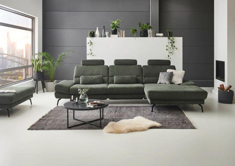 Modernes grünes Ecksofa mit verstellbaren Kopfstützen vor grauer Akzentwand mit weißem Wandregal in minimalistischem Wohnzimmer mit großem Fenster.