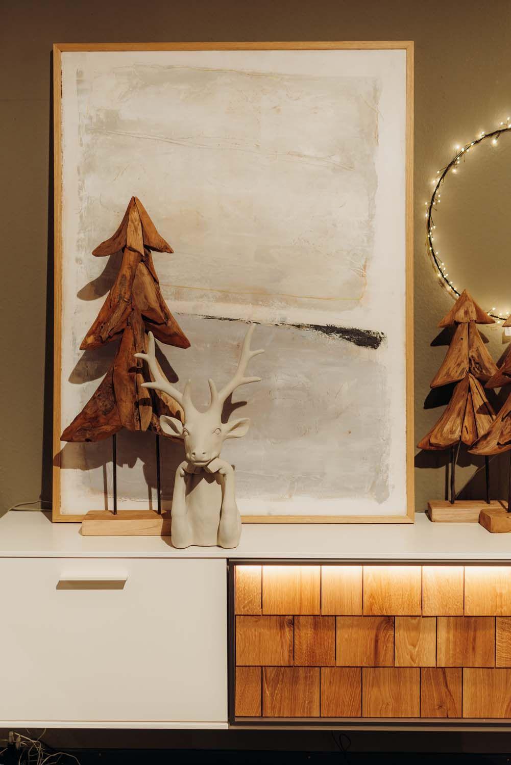 Detailaufnahme: Rustikale Holztannenbäume vor abstrakter Wandkunst mit weißer Hirschfigur als stimmungsvolle Winterdeko.