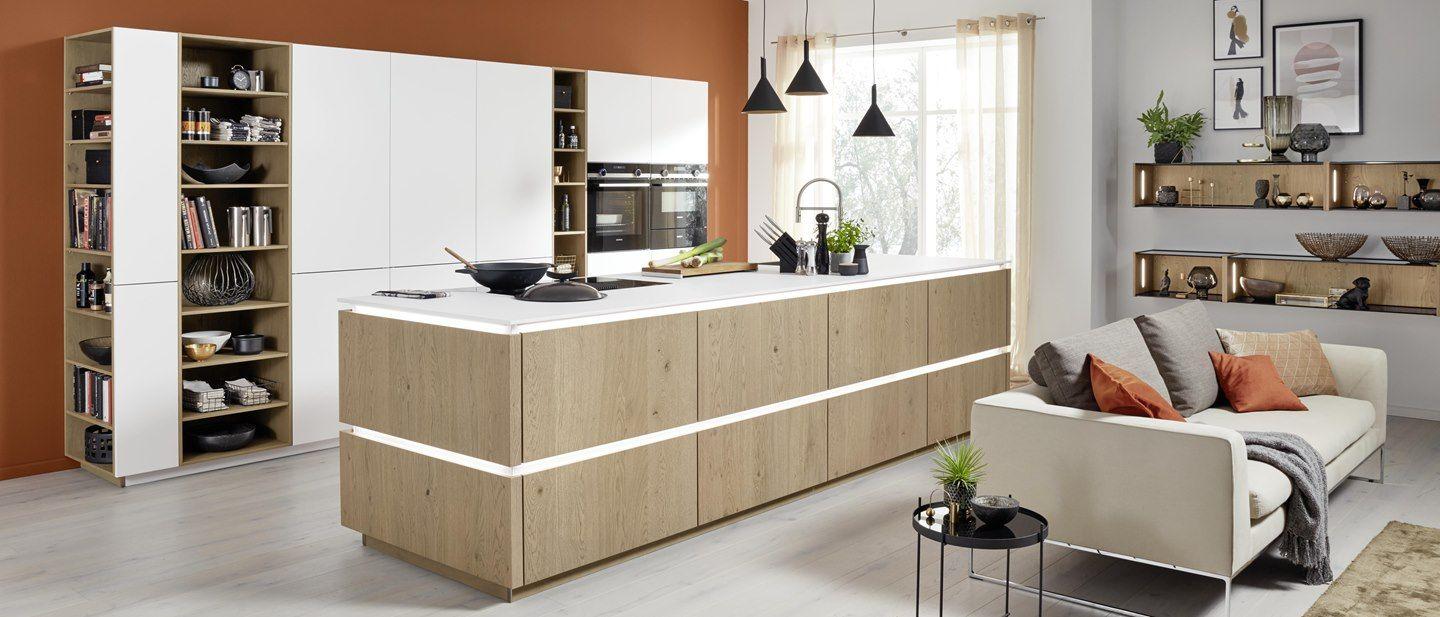 Große Kücheninsel in Eiche mit weißer Arbeitsplatte, LED-Beleuchtung, offenen Regalsystemen und angrenzendem Wohnbereich mit beiger Couch und Terrakotta-Akzentwand.