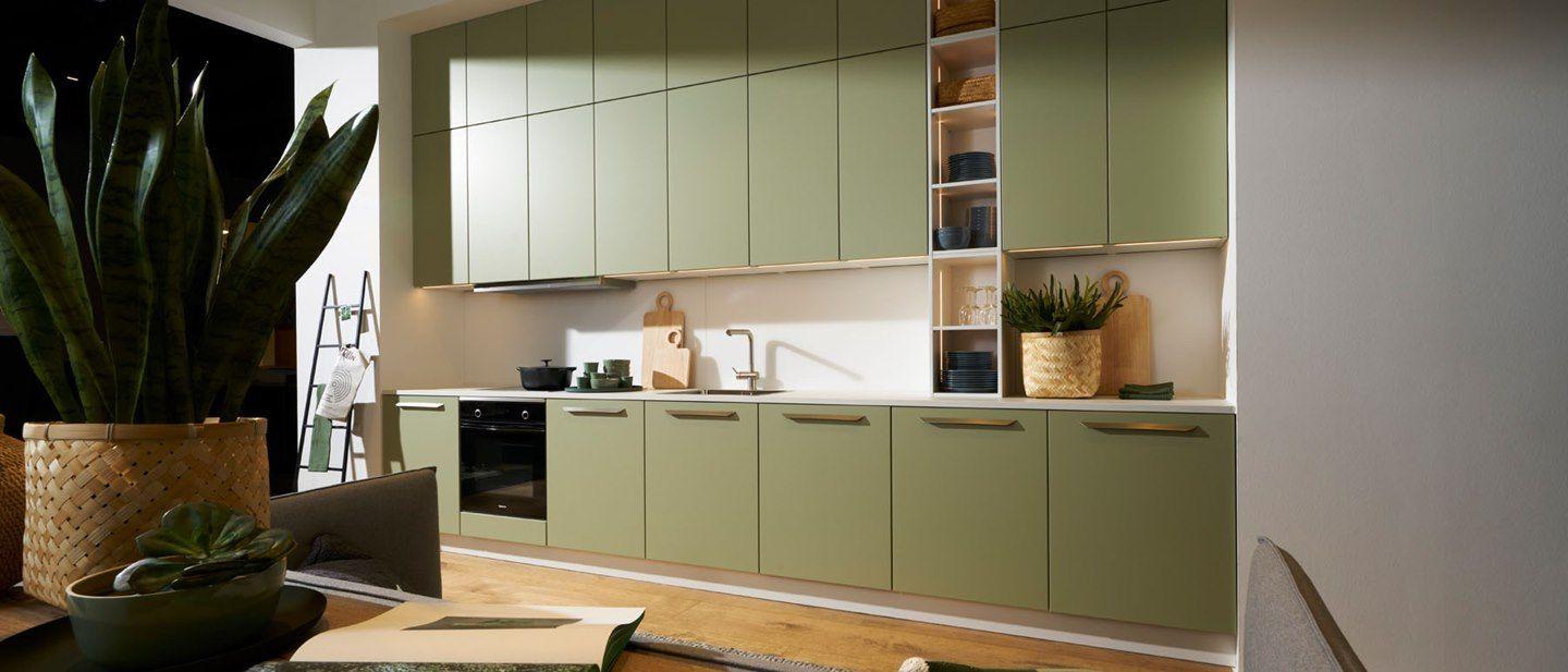 Moderne Küchenzeile in sanftem Grün mit eleganten Griffen, großzügigem Stauraum und offenen Regalen. Pflanzendekoration und Holzregale ergänzen das natürliche, warme Ambiente.
