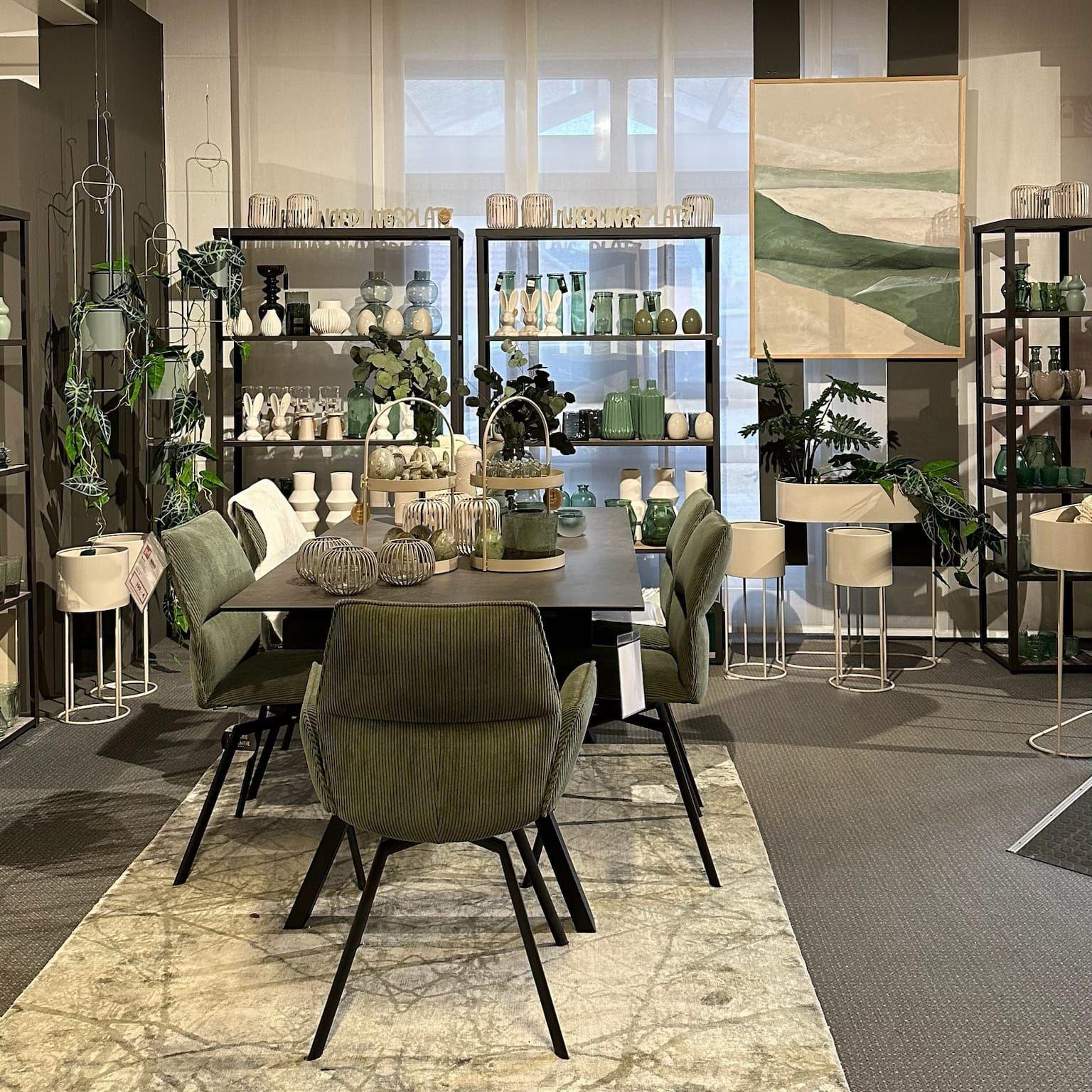 Moderne Essgruppe mit grünen Stühlen und schwarzem Tisch, umgeben von Pflanzen und grüner Glasdeko im Regal.