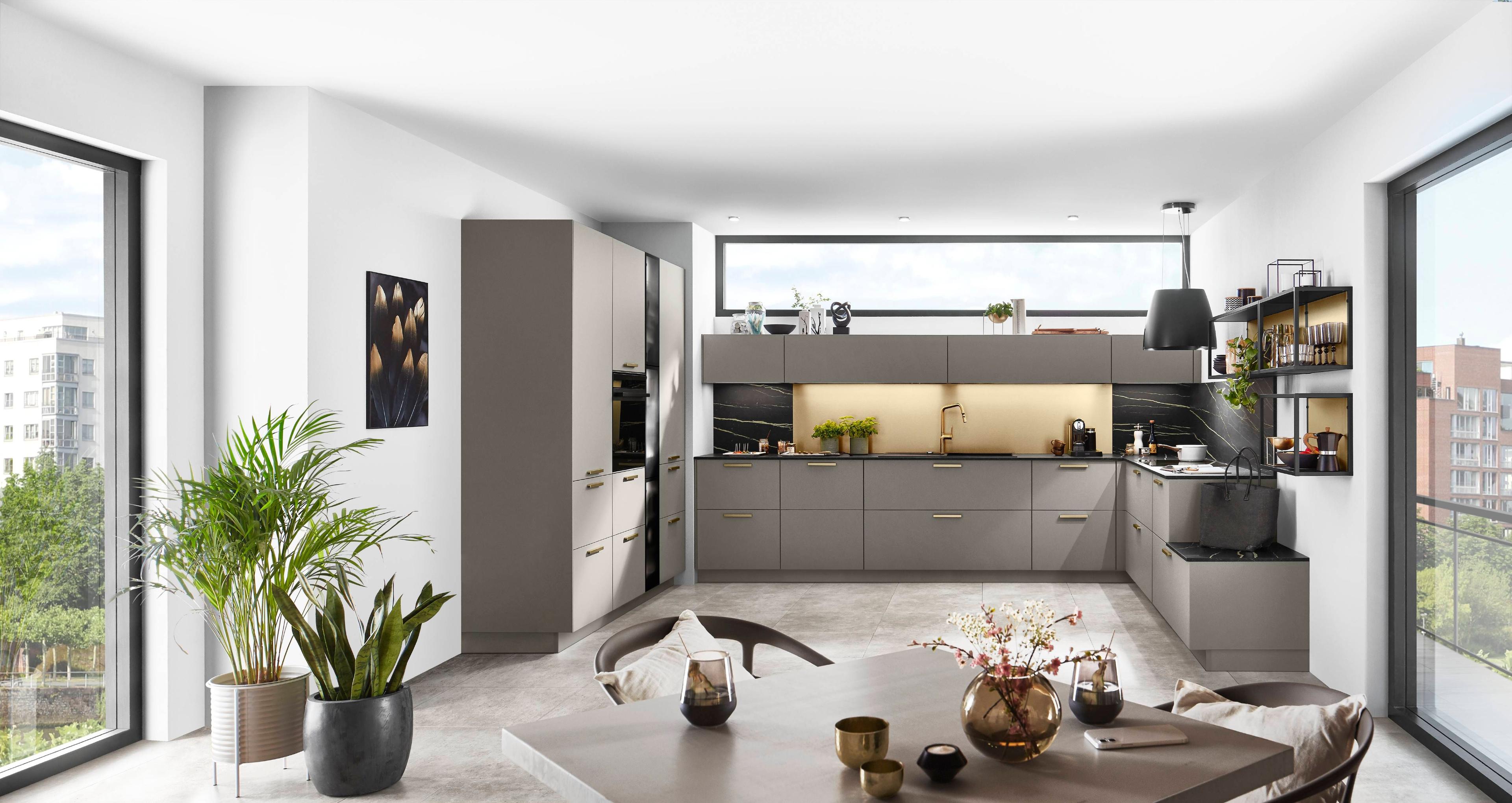 Moderne, offene Küche mit grauen Fronten, schwarzen Akzenten und goldenen Griffen, ergänzt durch große Fenster, Pflanzen und stilvolle Dekoration auf Regalen