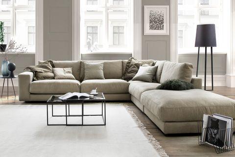 Großzügiges Ecksofa in Beige mit dekorativen Kissen in einem stilvollen, hellen Wohnraum mit Holzfußboden, hohen Fenstern und modernen Accessoires.
