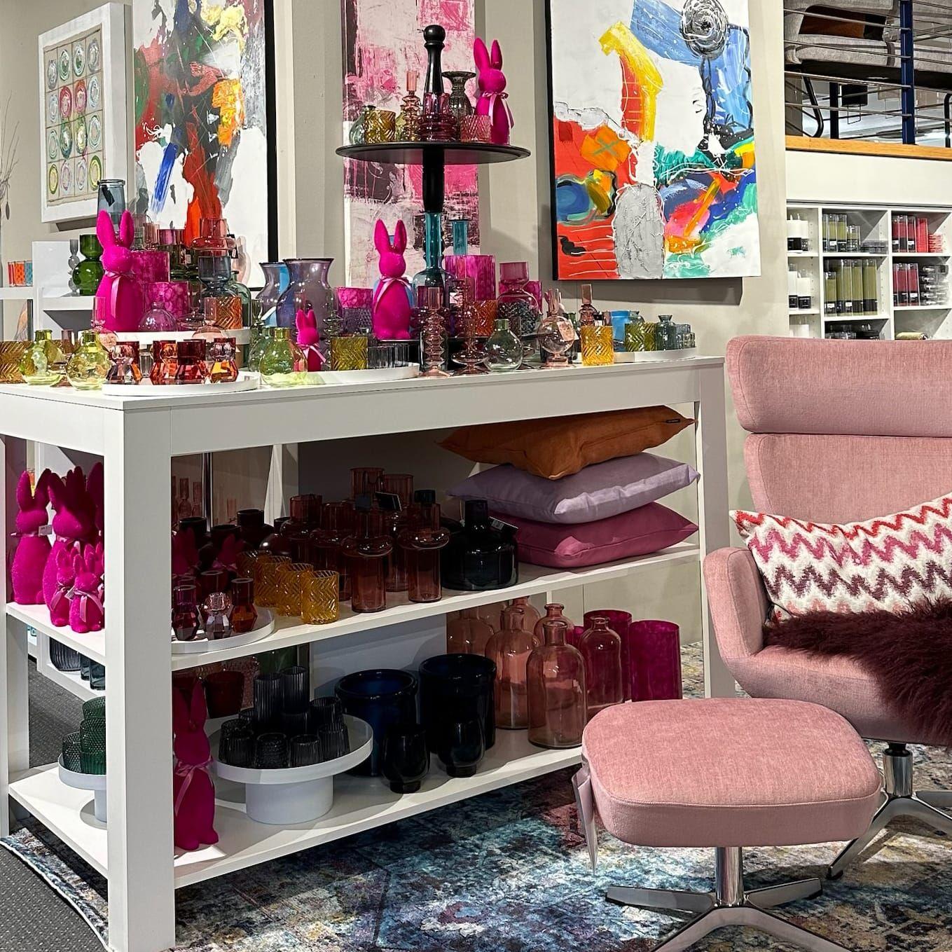 Bunte Boutique-Ausstellung mit pinken Osterhasen, farbigen Glasvasen und abstrakten Kunstwerken neben rosafarbenem Sessel.
