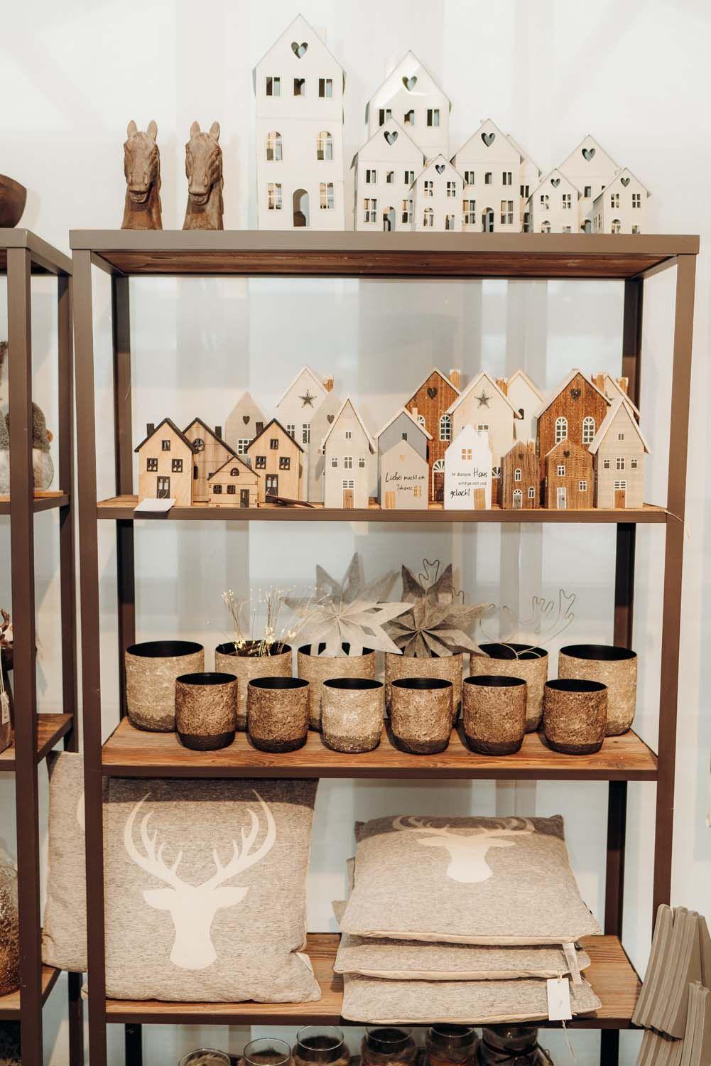 Holzregal mit weißen Deko-Häuschen, Hirschfiguren, goldenen Windlichtern und natürlichen Textilien in warmer Atmosphäre.