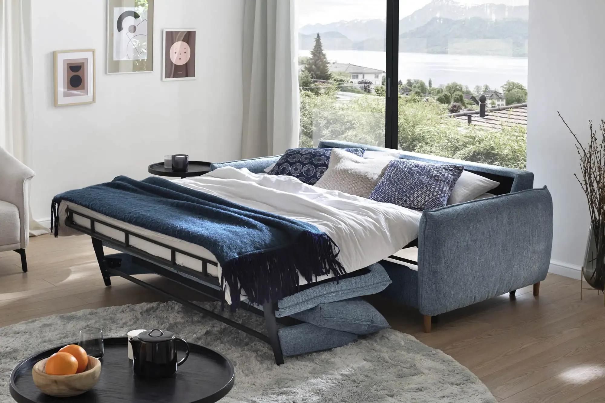Ausgeklapptes Schlafsofa in Blau mit Decke und Kissen, aufgestellt in einem hellen Zimmer mit Panoramablick – funktional und einladend zugleich.