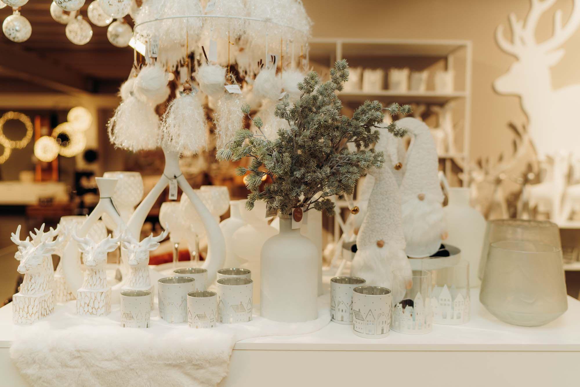 Festliche Winterdeko mit weißen Figuren, Tannenzweigen in moderner Vase und stilvollen Windlichtern in heller Präsentation.
