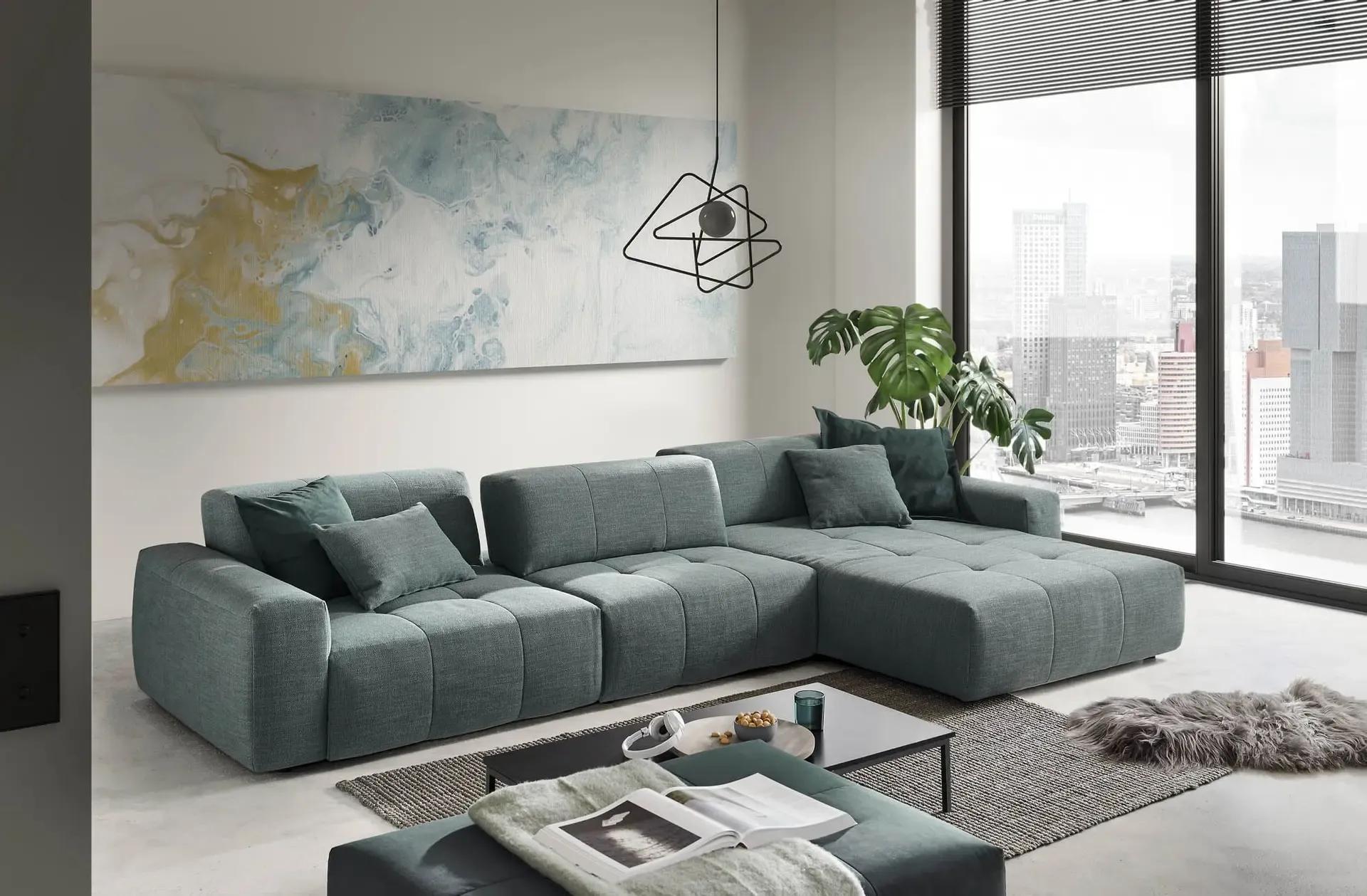 Graues Modulsofa mit Chaiselongue vor Panoramafenster mit Stadtblick, kombiniert mit abstraktem Wandbild und geometrischer Pendelleuchte.