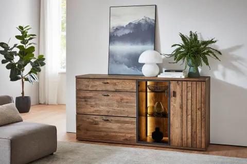 Rustikale Kommode aus dunklem Holz mit drei Schubladen und beleuchteter Glastür – dekorativ inszeniert mit Lampe, Büchern und Zimmerpflanze.