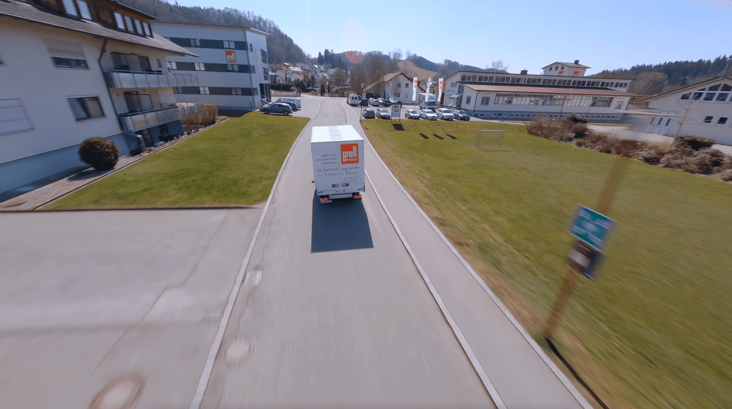Drohnenaufnahme eines weißen Lieferwagens mit rotem Logo auf Landstraße durch ein Gewerbegebiet mit modernen Gebäuden und bewaldeten Hügeln im Hintergrund.