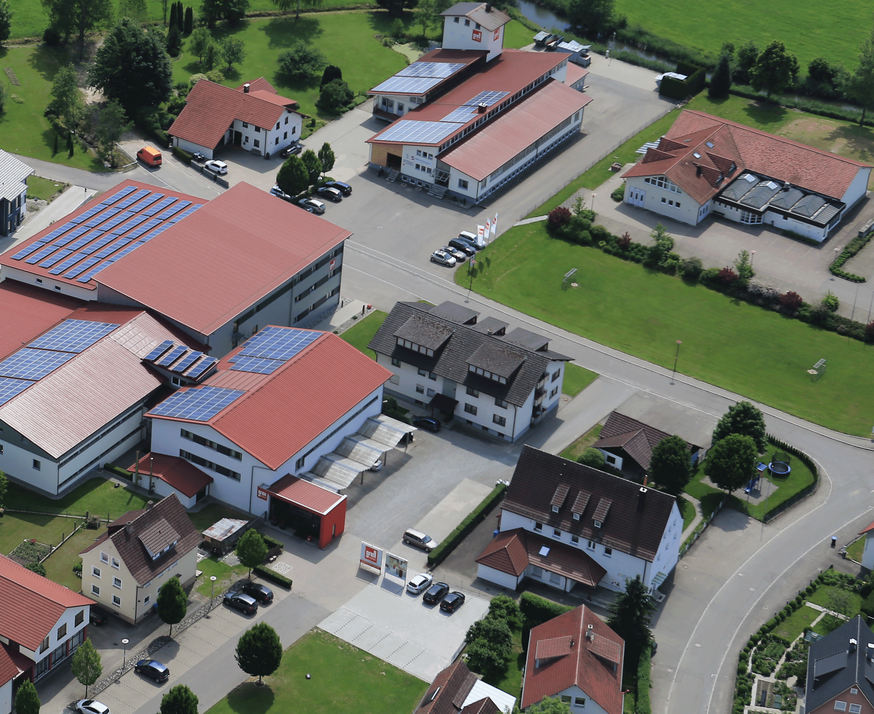 Luftaufnahme eines Industriekomplexes mit mehreren Gebäuden mit roten Dächern und Solaranlagen, umgeben von grünen Feldern und Wohnhäusern in ländlicher Umgebung.