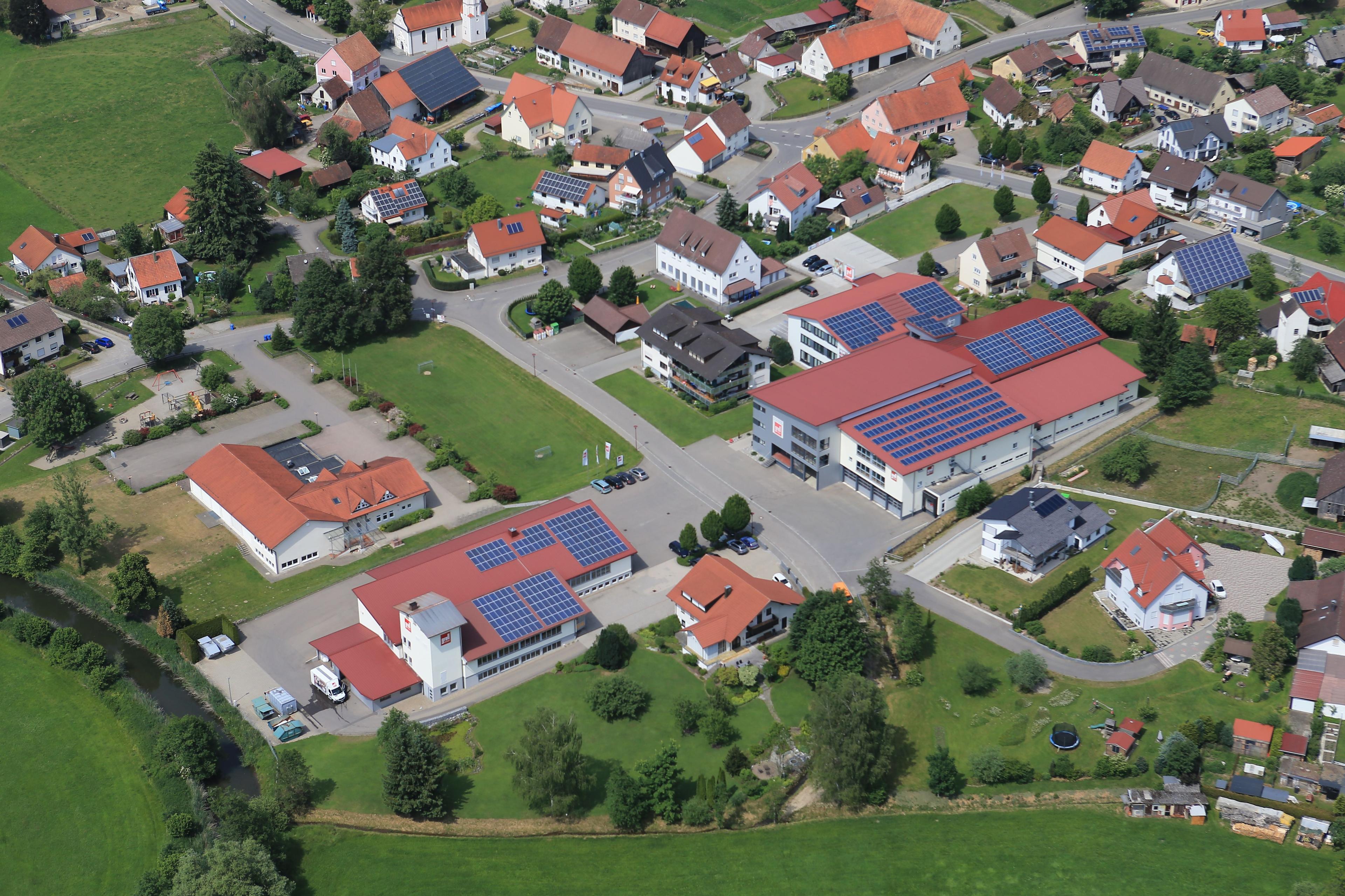 Luftaufnahme eines ländlichen Gewerbegebiets mit modernen Industriegebäuden mit Solaranlagen auf roten Dächern, umgeben von traditionellen Wohnhäusern und grünen Feldern.