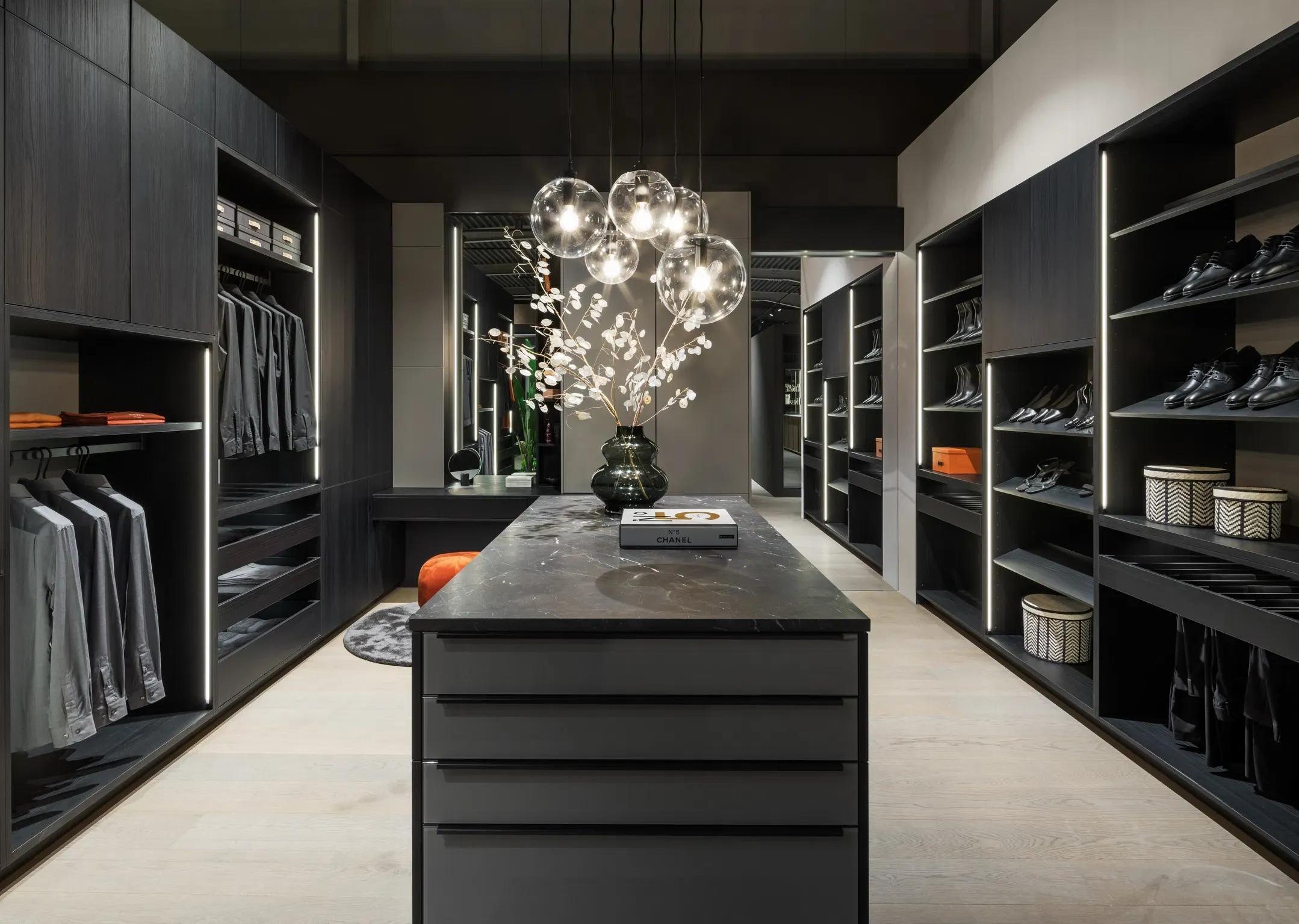 Elegantes Ankleidezimmer in dunkler Holzoptik mit zentraler Inselkommode, offener Schranklösung und stilvoller Beleuchtung.