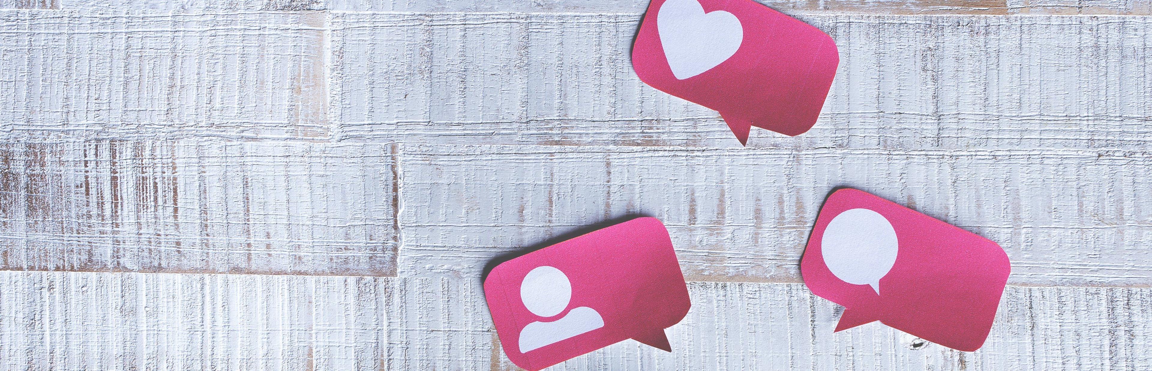 Drei rosa Social-Media-Symbole auf weißem Holzhintergrund zeigen Like-Herz, Nutzer-Profil und Sprechblase für digitale Kommunikation und Interaktion.