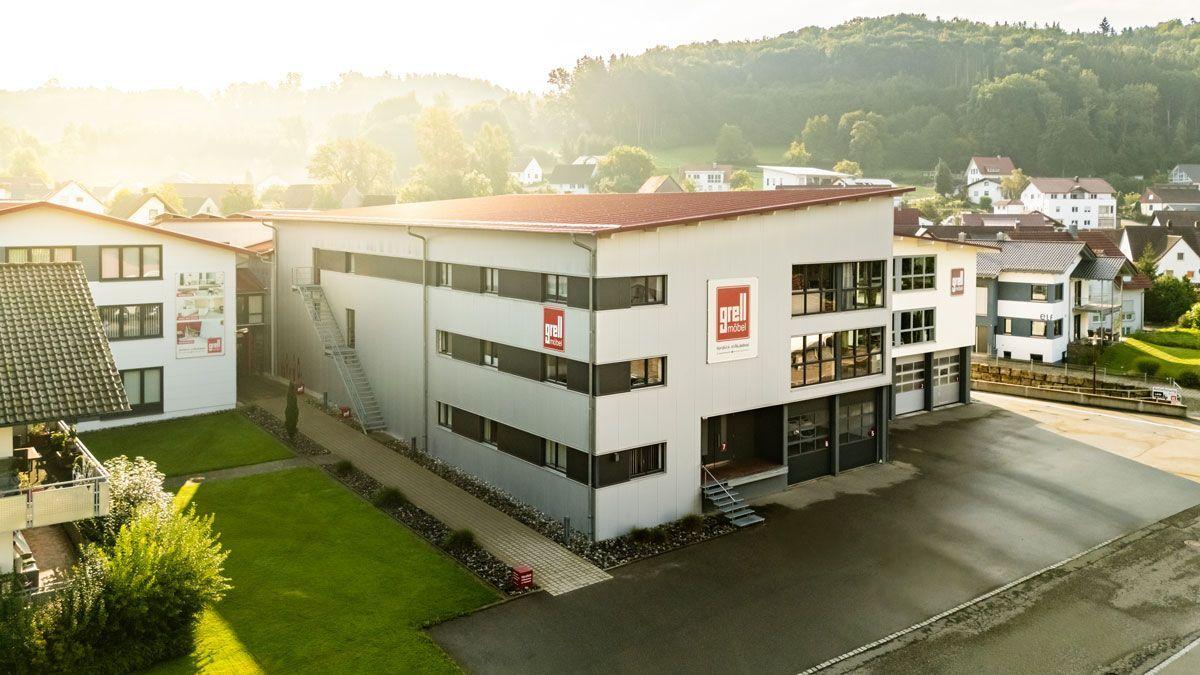 Nahaufnahme eines modernen dreistöckigen weißen Gewerbegebäudes mit rotem Dach und roten Firmenschildern, umgeben von Rasenflächen und bewaldeten Hügeln.