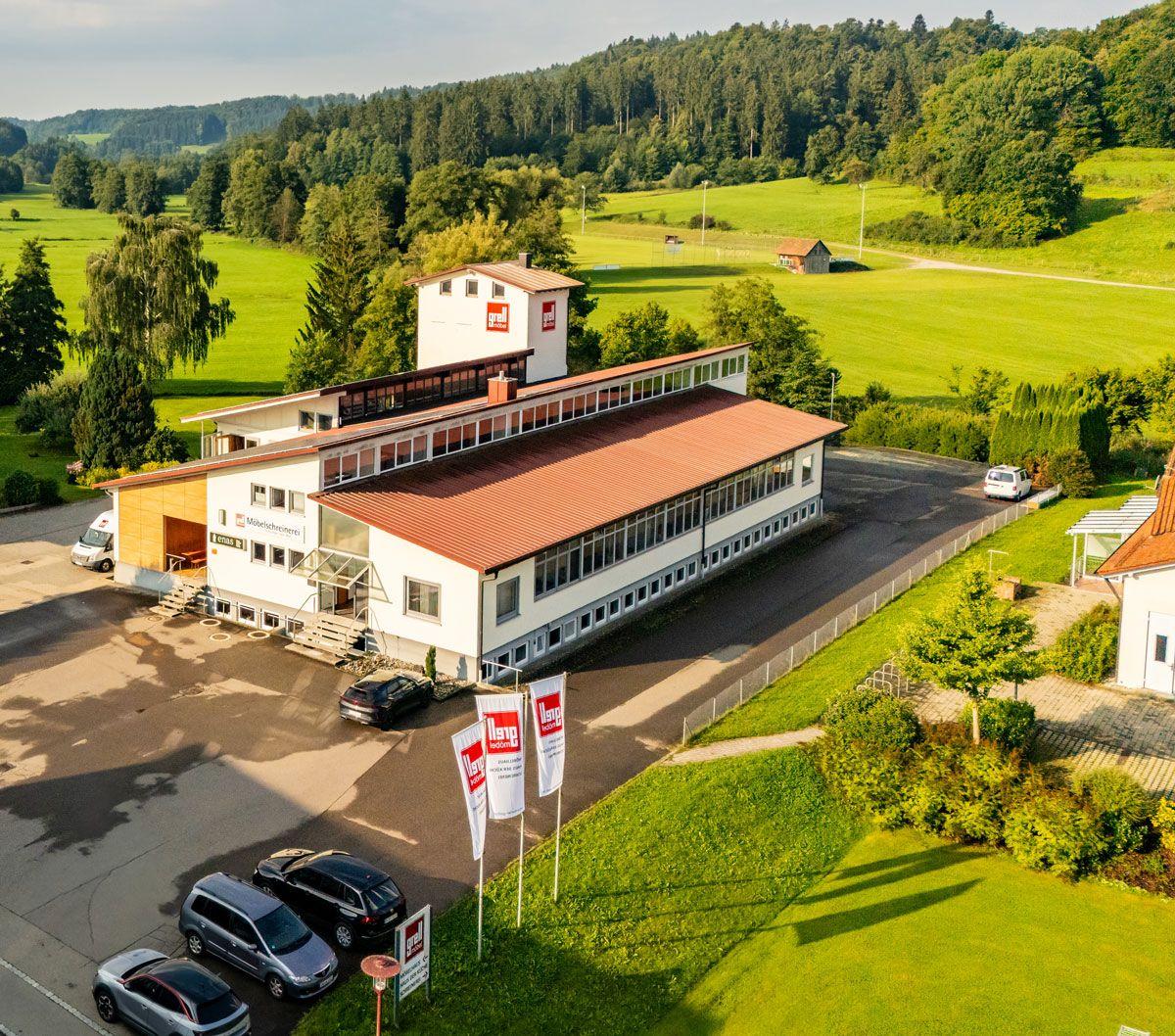 Luftaufnahme eines modernen Gewerbegebiets mit weißen Bürogebäuden und Industrieanlagen, eingebettet in eine grüne Hügellandschaft mit einer Wohnsiedlung im Hintergrund bei warmem Sonnenlicht.