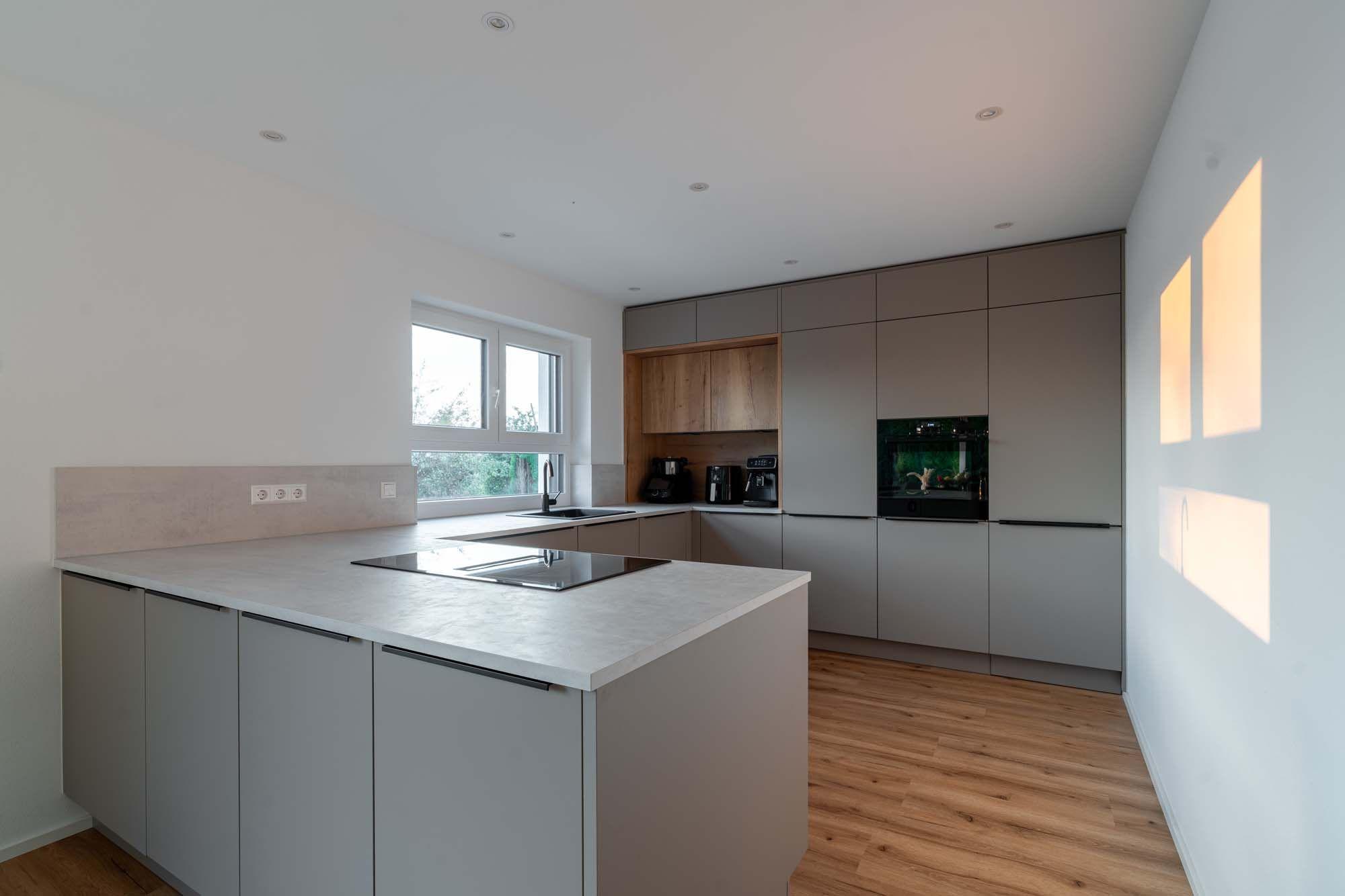 Moderne Küche mit grauer Kücheninsel, beigen Fronten und Holzelementen, Blick durchs Fenster auf Grünfläche