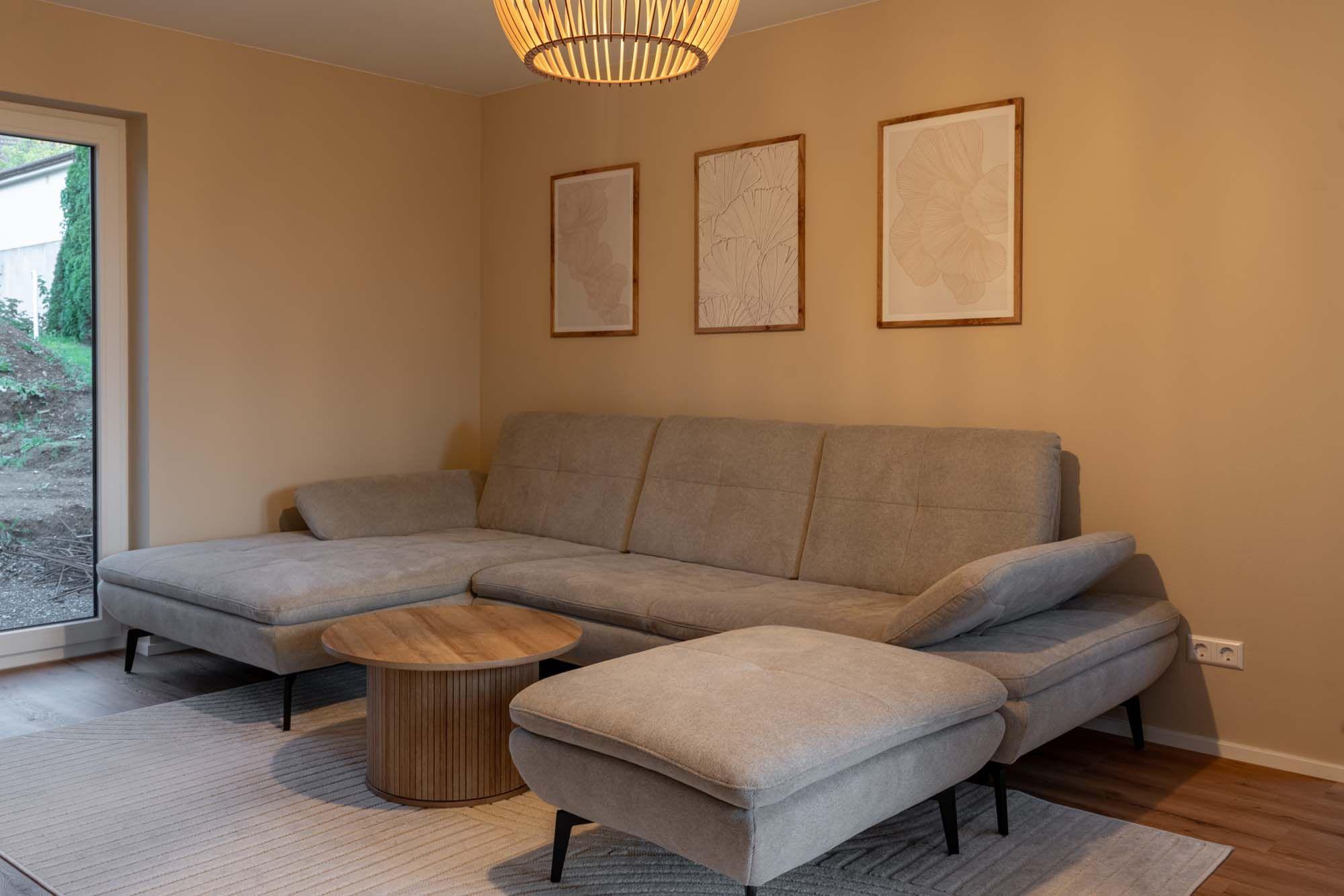 Wohnzimmer mit großer beiger Wohnlandschaft, rundem Holzcouchtisch und drei gerahmten Wandbildern.