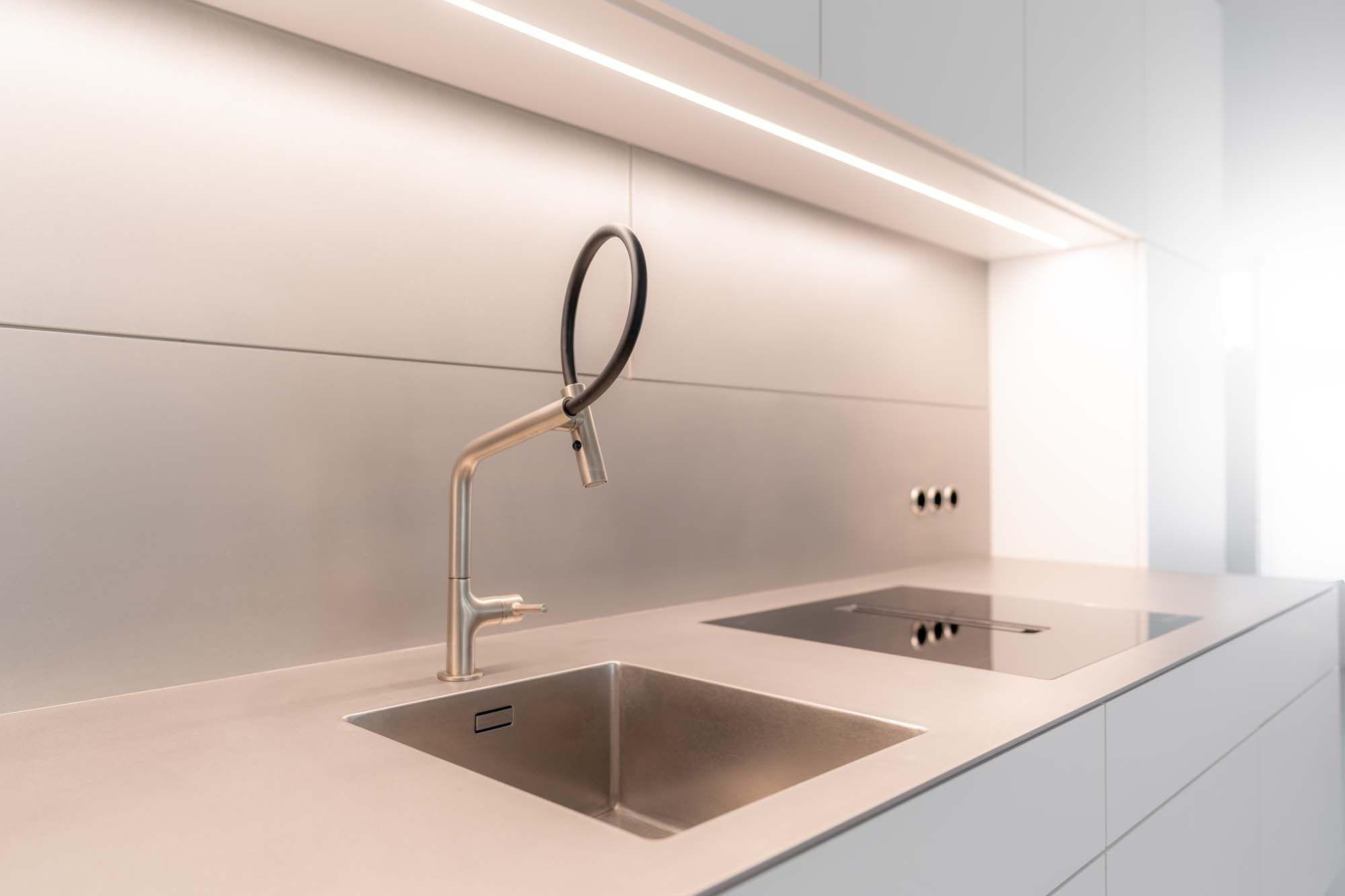 Nahaufnahme einer Edelstahlspüle mit geschwungenem Chrom-Wasserhahn vor beleuchteter Rückwand.