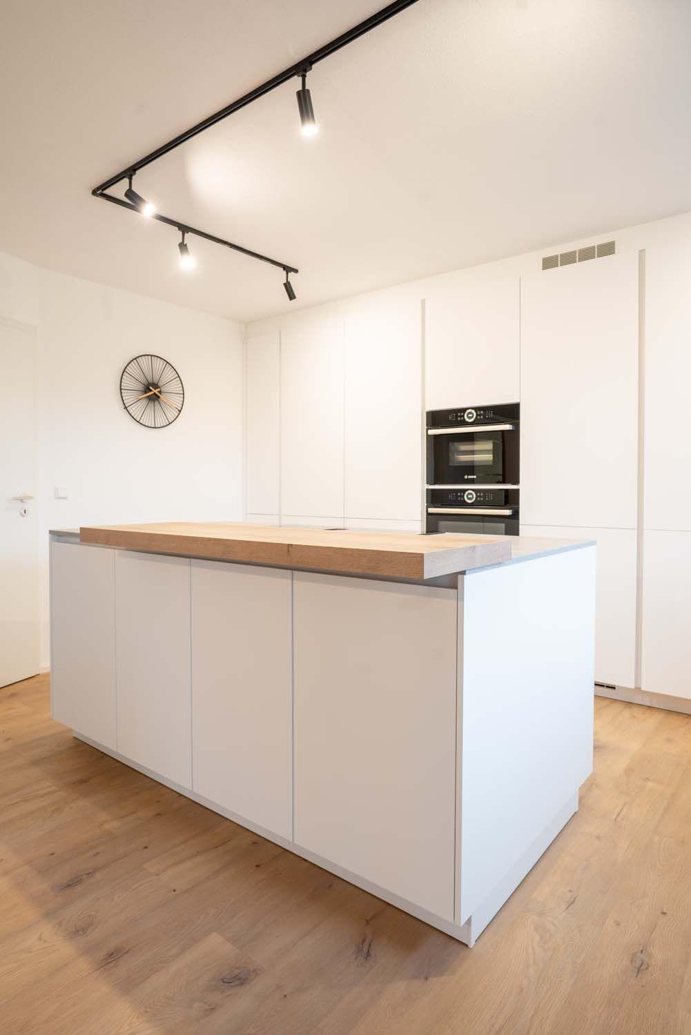 Weiße Kochinsel mit Holzarbeitsplatte vor deckenhoher Gerätewand mit schwarzen Einbaugeräten, Design-Wanduhr und schwarzem Strahler-System.
