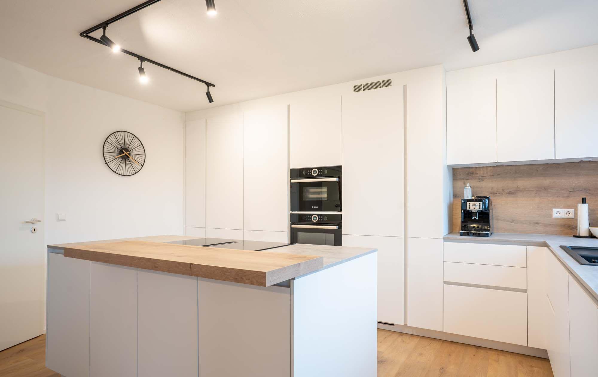 Moderne Küche mit Kochinsel aus Holz und grauer Arbeitsplatte, weißen Hochschränken mit schwarzen Geräten und Holz-Rückwand im Spülbereich.