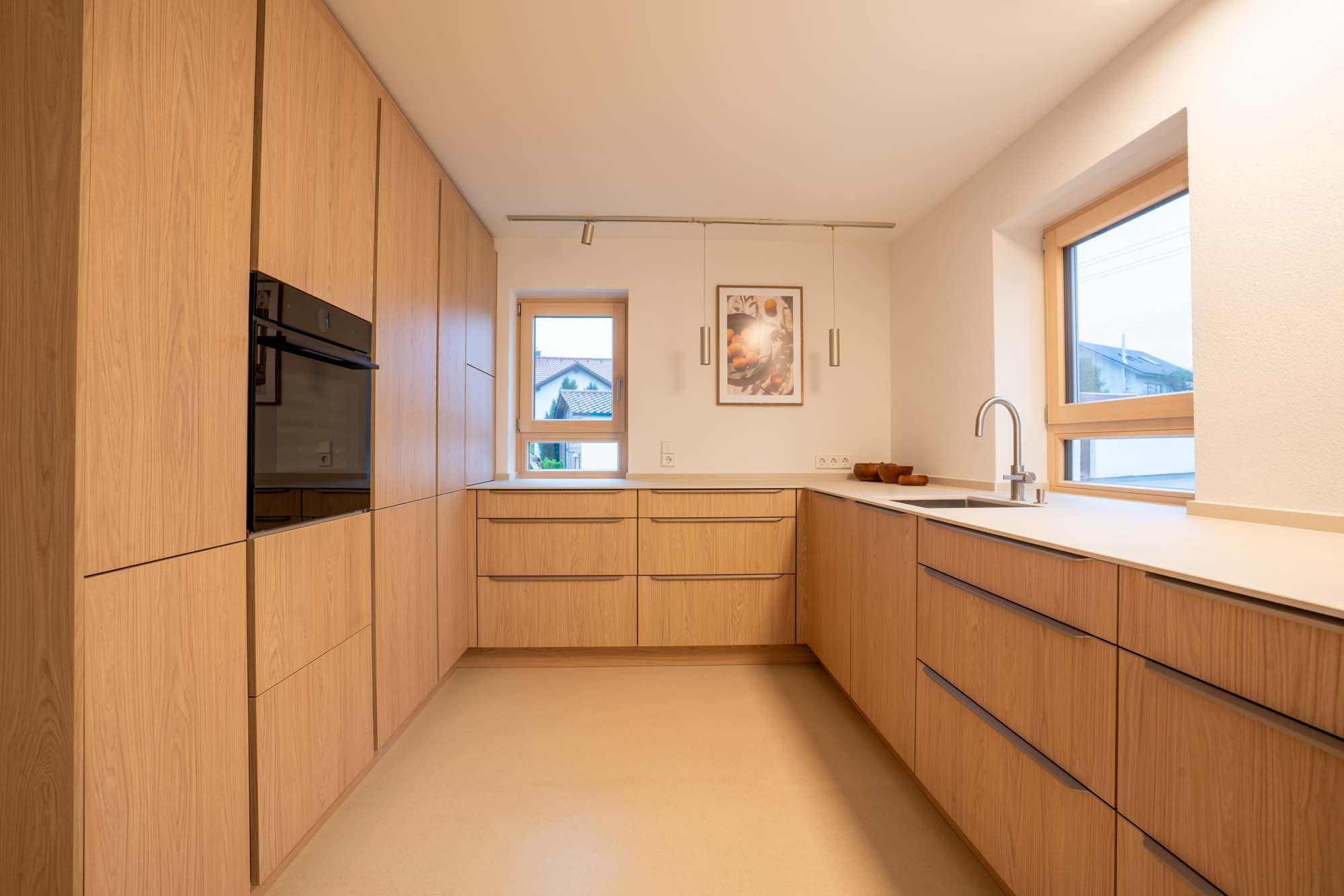 Moderne Küche komplett aus Eiche mit eingebautem Backofen, beigem Boden und mittig platziertem Fenster.