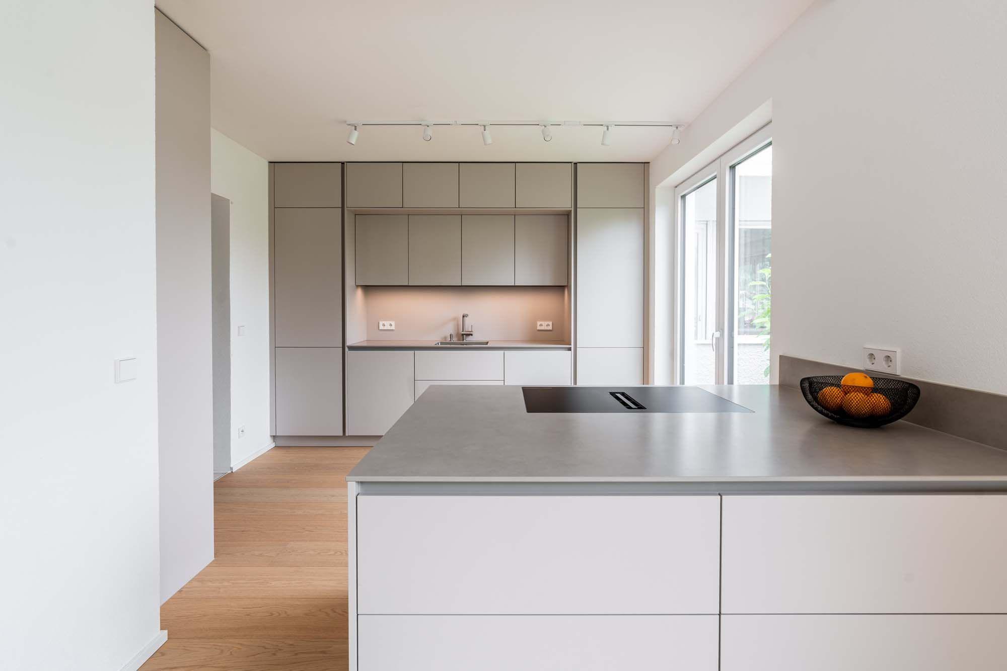 Offene Küche mit weißer Kochinsel, beige-taupefarbenen Hochschränken, LED-Schienenbeleuchtung und hellem Holzboden.