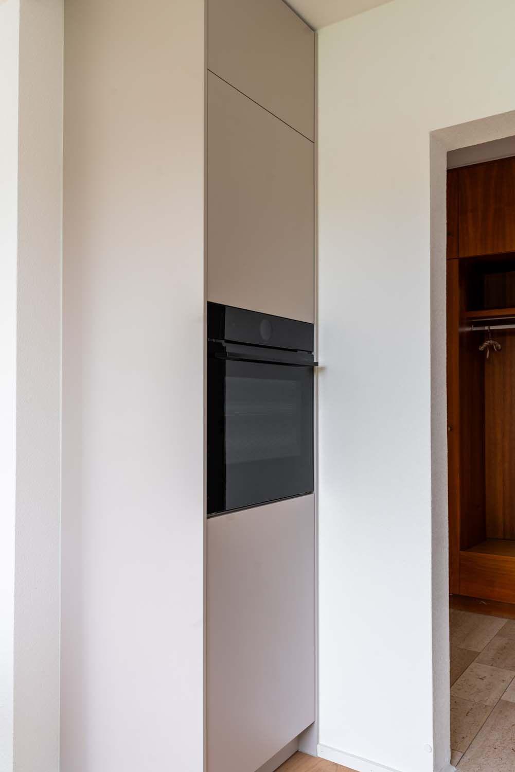 Eingebauter schwarzer Backofen in beige-weißer Hochschrankfront, Durchgang zur Garderobe mit Holzelementen.