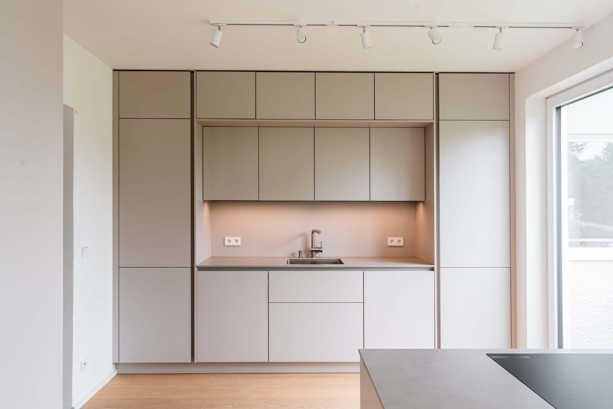 Frontalansicht moderne Einbauküche mit symmetrischer Schrankaufteilung in Beige und Weiß, LED-Schienenbeleuchtung und Kochinsel.