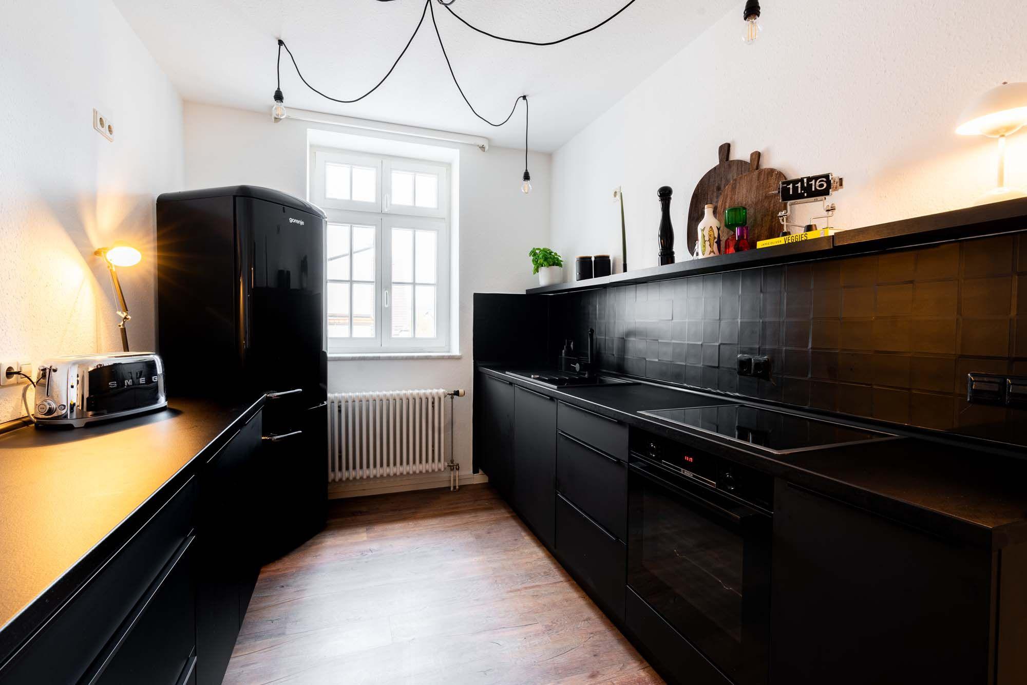 Zweizeilige schwarze Küche mit glänzendem Retro-Kühlschrank, schwarzen Mattfronten, Fliesenwand, offenem Wandregal mit Holzbrettern, hängenden Glühbirnen-Pendelleuchten und hellem Holzboden.