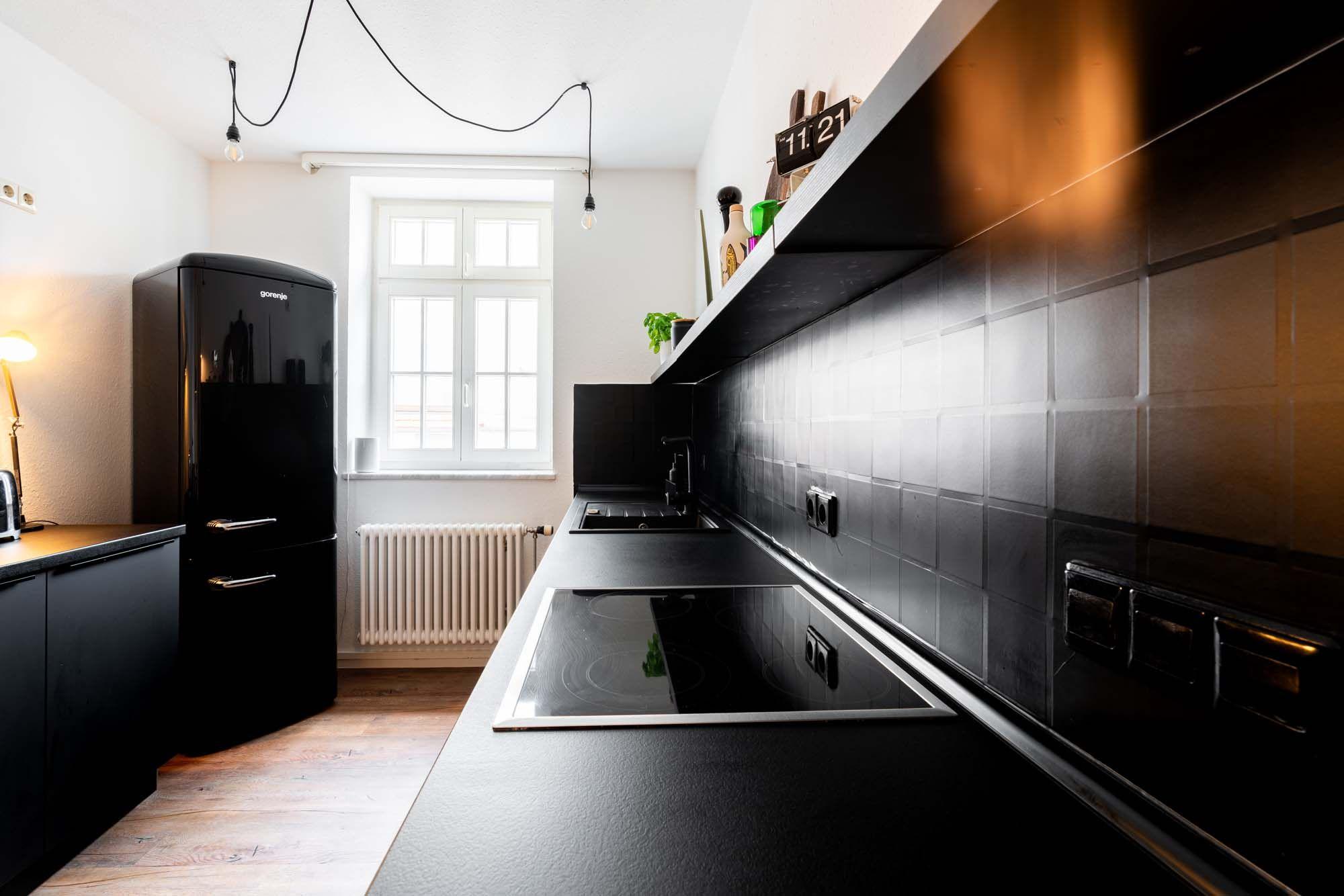 Schwarze Küchenarbeitsplatte mit Induktionskochfeld, glänzender Fliesenwand, offenem Wandregal, Retro-Kühlschrank im Hintergrund und geschwungenen Pendelkabeln.