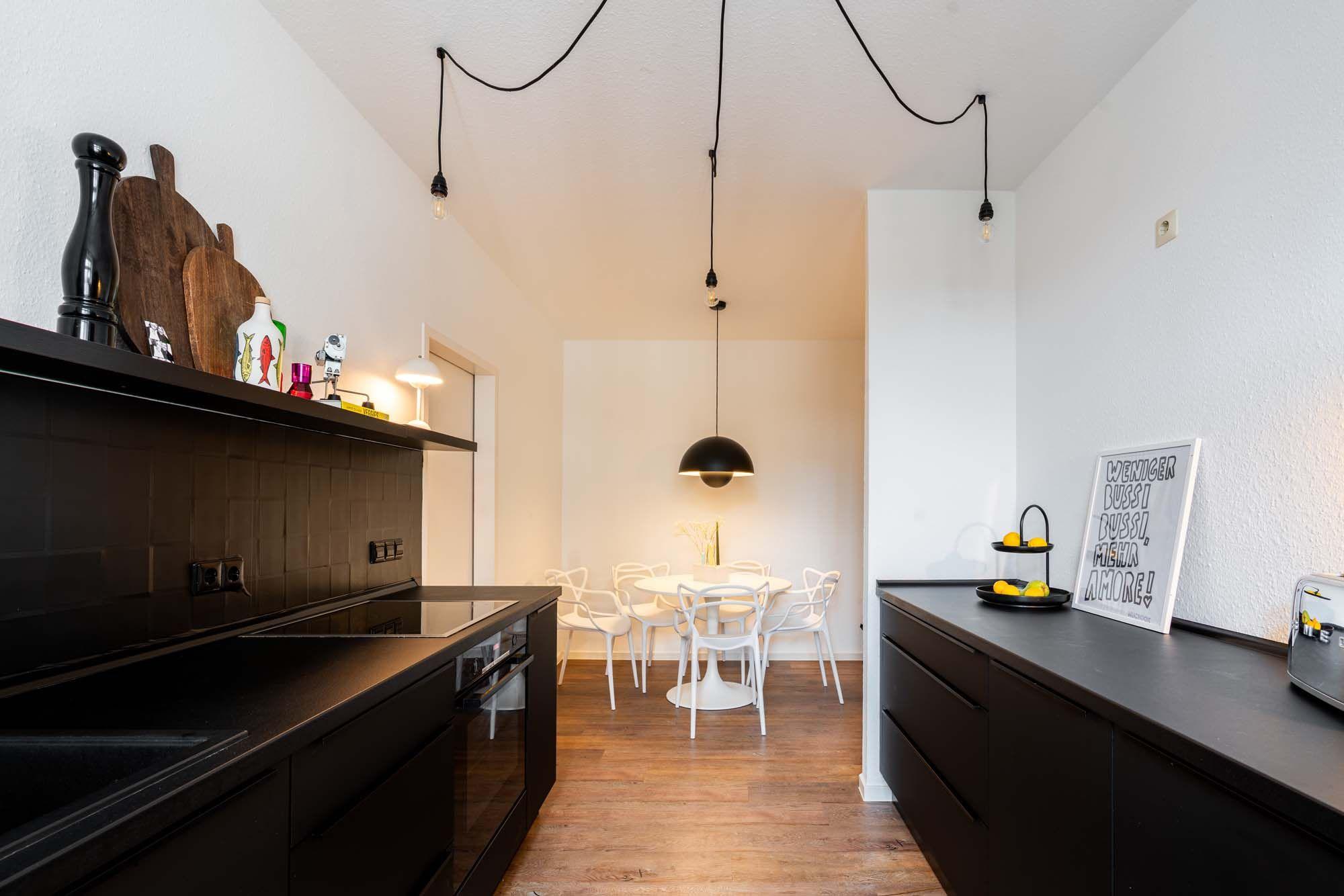 Blick von schwarzer Küche mit Holzregal-Deko in offenen Essbereich mit weißem Tulip-Tisch, Design-Stühlen, schwarzer Halbkugel-Pendelleuchte und Typografie-Poster.