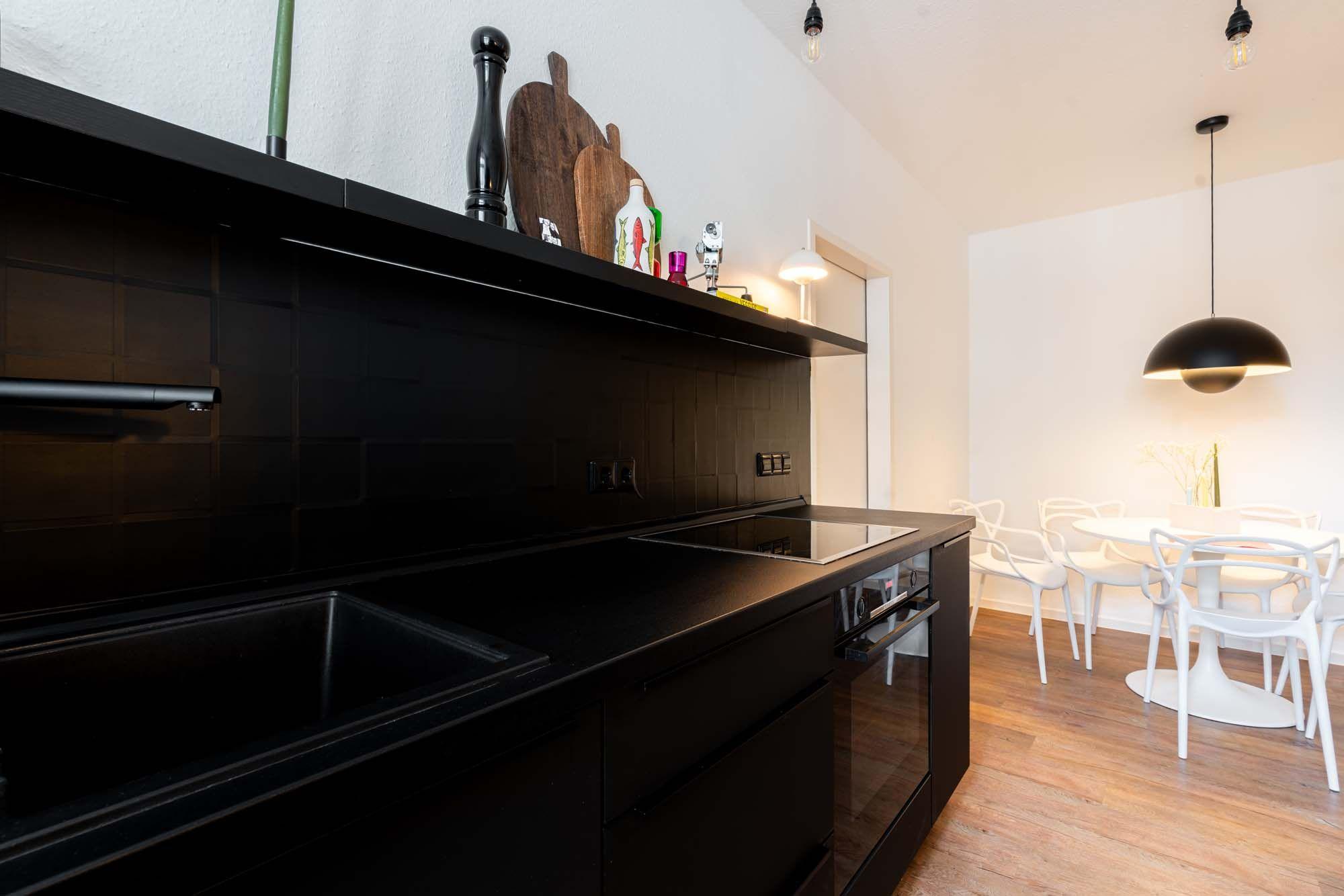 Nahaufnahme schwarze Küche mit offenem Wandregal, bunten Deko-Objekten, Holz-Fliesenwand, matter Arbeitsplatte, Induktionskochfeld und Durchblick zum weißen Essbereich mit Pendelleuchte.