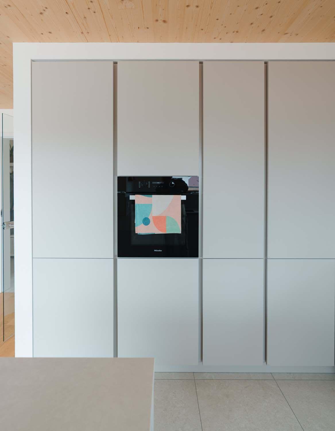 Grifflose Hochschrank-Front mit mittig eingebautem schwarzem Backofen unter Holzdecke.