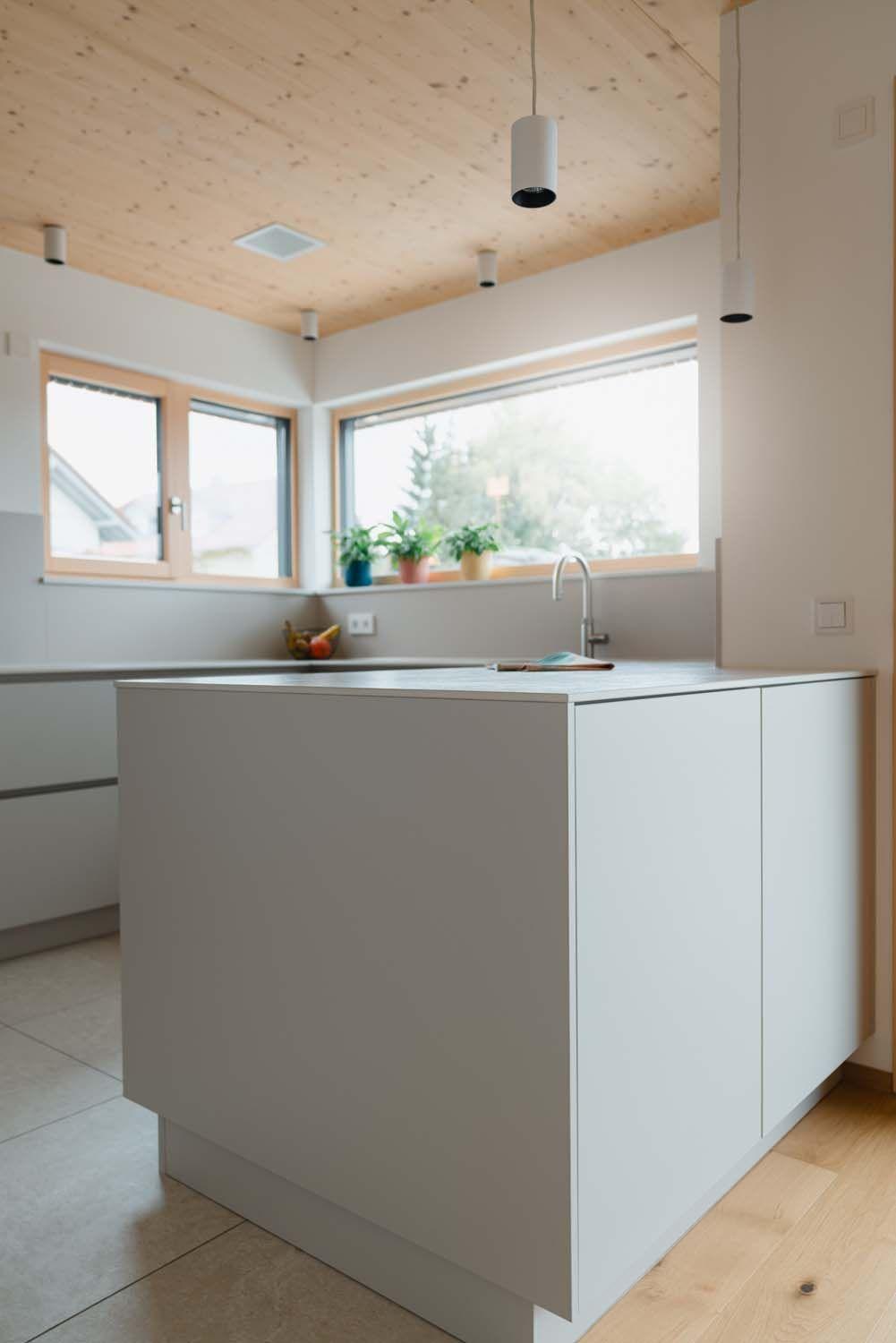 Minimalistische Kücheninsel in Hellgrau vor großem Fenster mit Holzrahmen und natürlichem Lichteinfall.