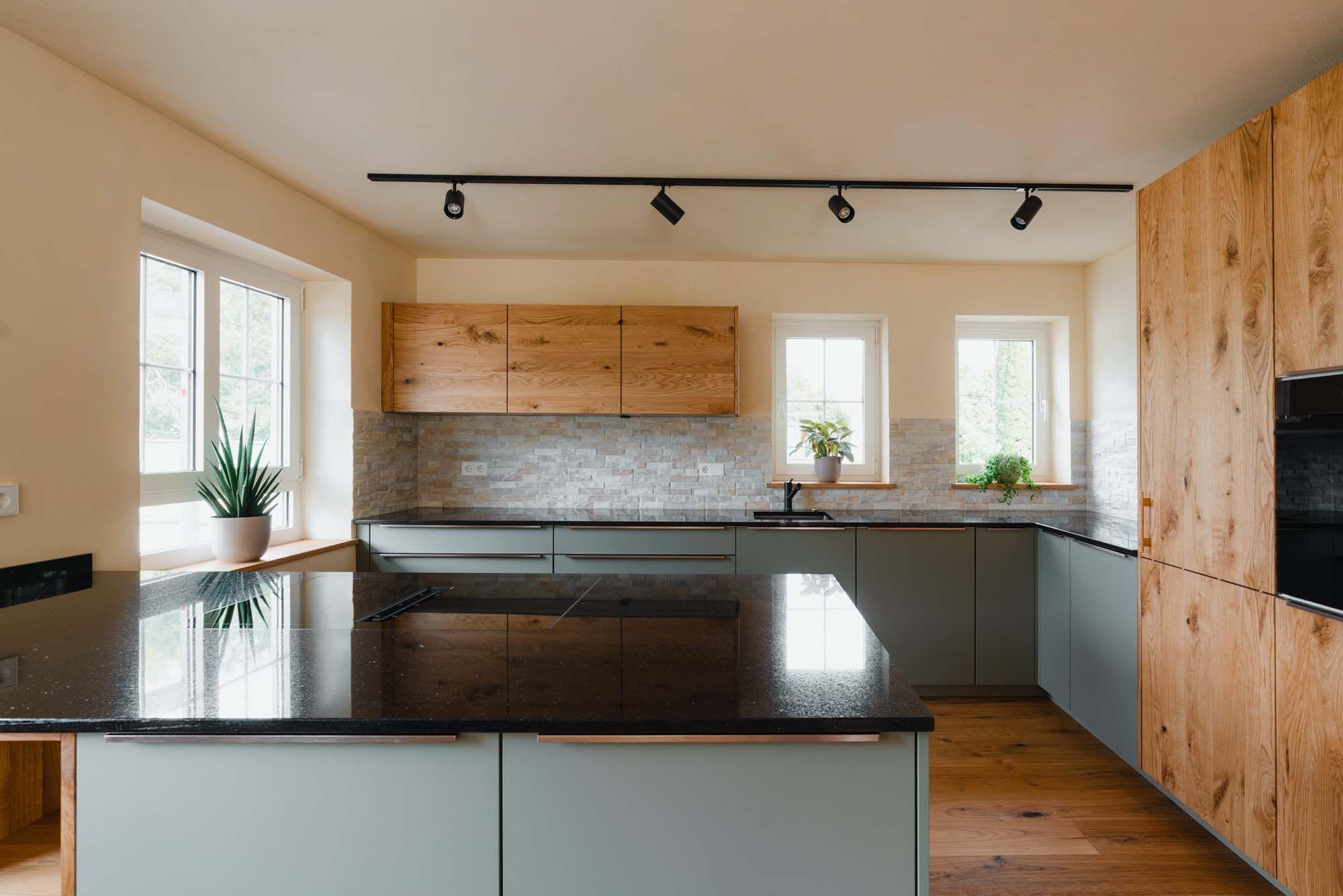 Offene Wohnküche mit salbeigrüner Kochinsel, schwarzer Granitarbeitsplatte, Holz-Oberschränken, Steinverblender-Rückwand, schwarzem Track-Lighting und hellem Eichenboden.
