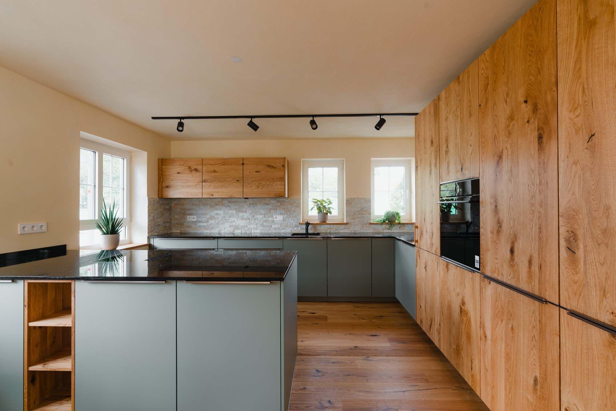 Moderne Küche mit salbeigrüner Insel mit offenem Holzregal, schwarzer Arbeitsplatte, Steinverblender-Rückwand und deckenhoher Holz-Gerätewand.