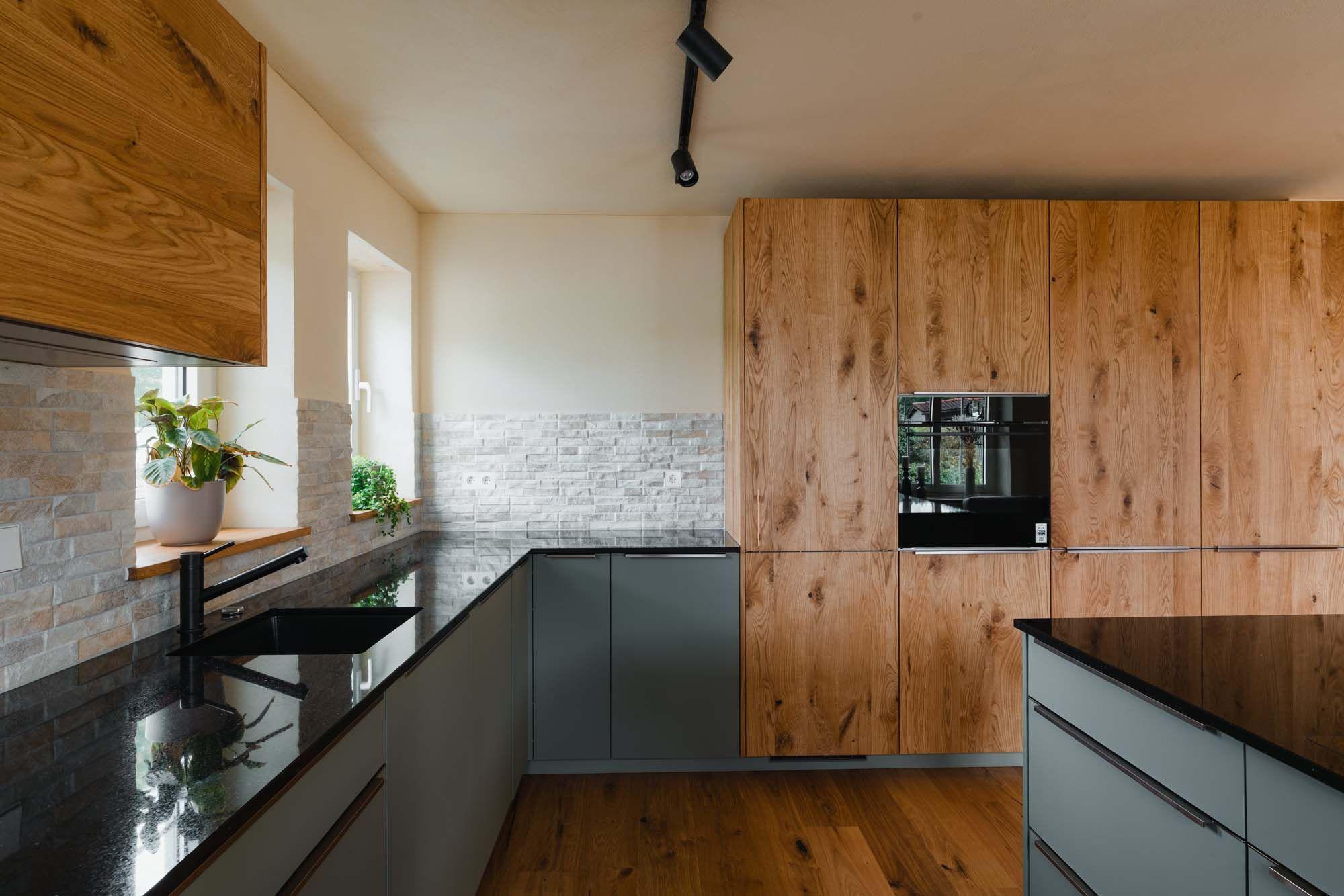 Kücheneckbereich mit salbeigrünen Unterschränken, schwarzer Granitarbeitsplatte, Steinverblender-Rückwand und hoher Holz-Hochschrankzeile mit integriertem schwarzem Backofen.v