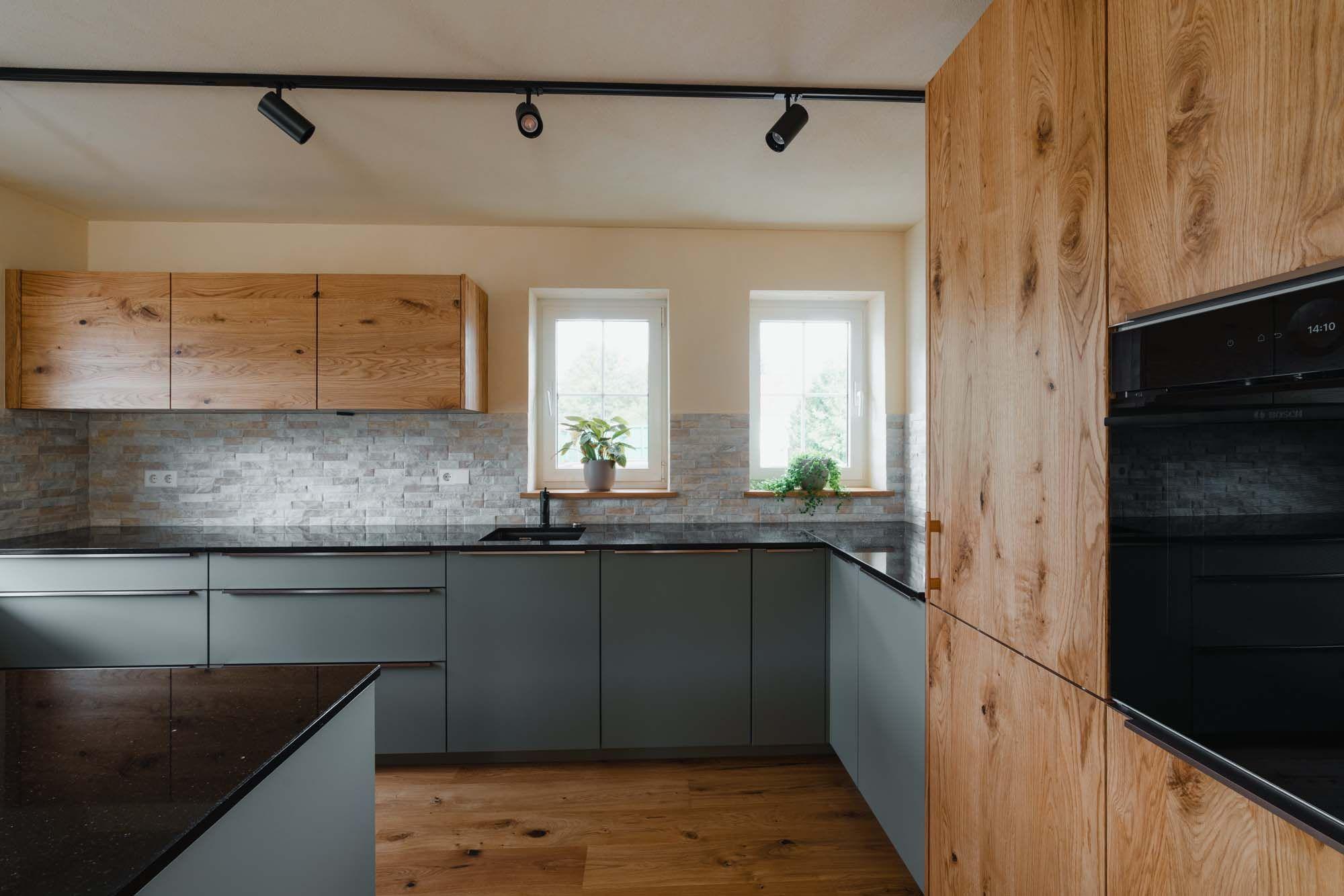 L-förmige Küchenzeile in Salbeigrün mit Holz-Oberschränken, Steinverblender-Rückwand, schwarzer Arbeitsplatte und zwei Fenstern mit Zimmerpflanzen.