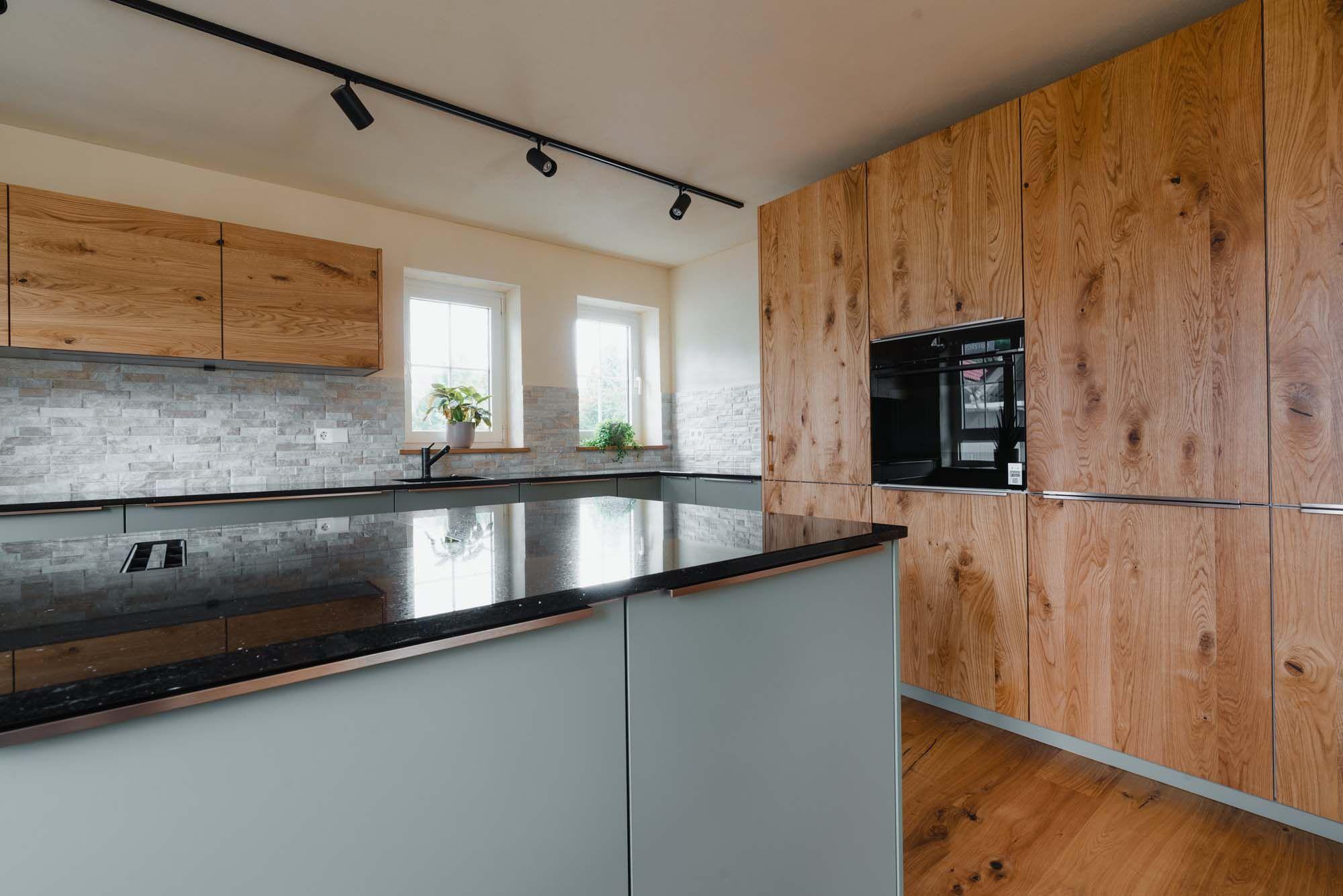 Offene Küche mit Kochinsel, salbeigrünen Fronten, schwarzer Granitarbeitsplatte, Steinverblender-Rückwand und Holz-Oberschränken unter schwarzer Schienenbeleuchtung.