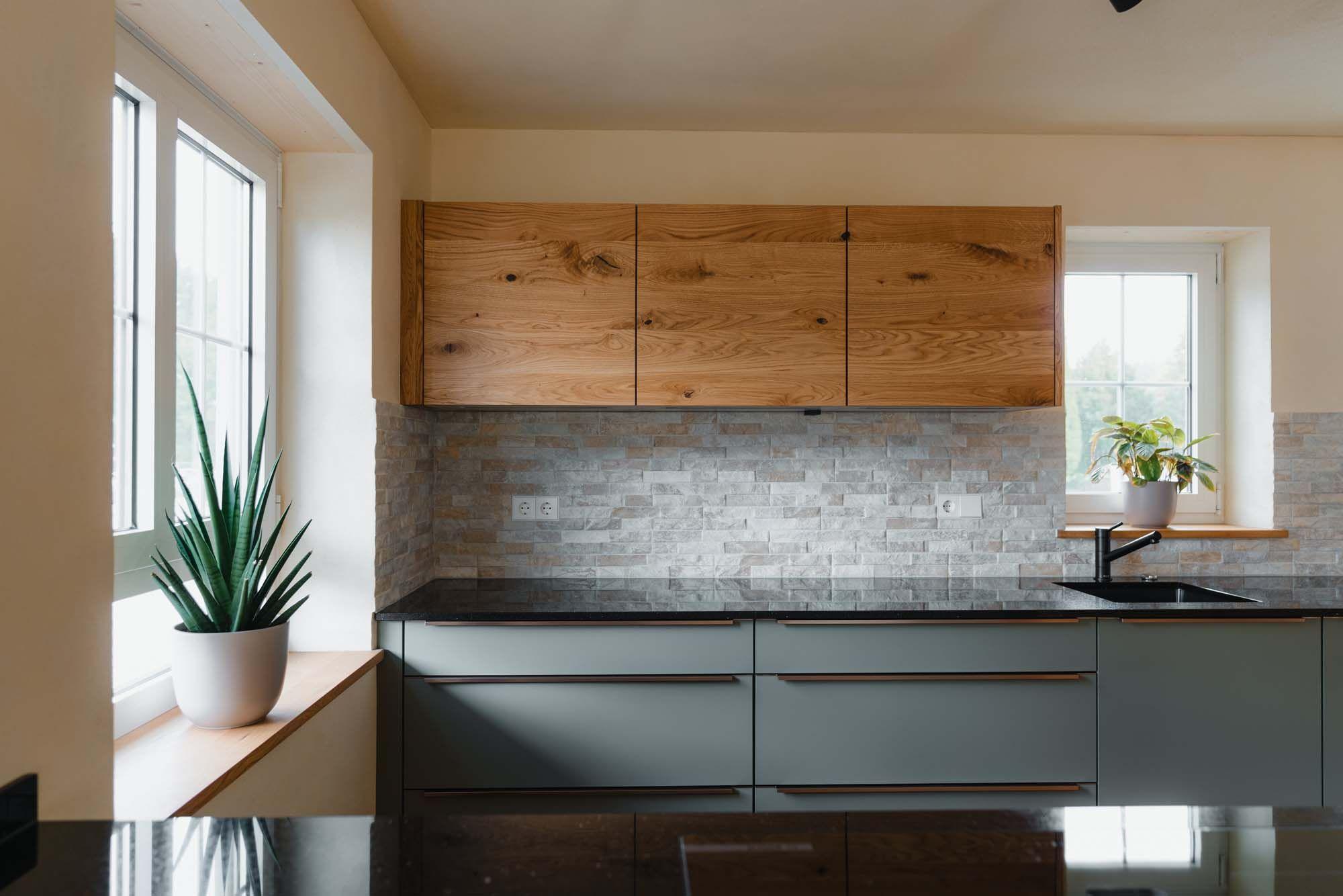 Küchenzeile mit Holz-Oberschränken, salbeigrünen Unterschränken, Steinverblender-Rückwand, großem Fenster links mit Aloe Vera und spiegelnder schwarzer Granitarbeitsplatte.