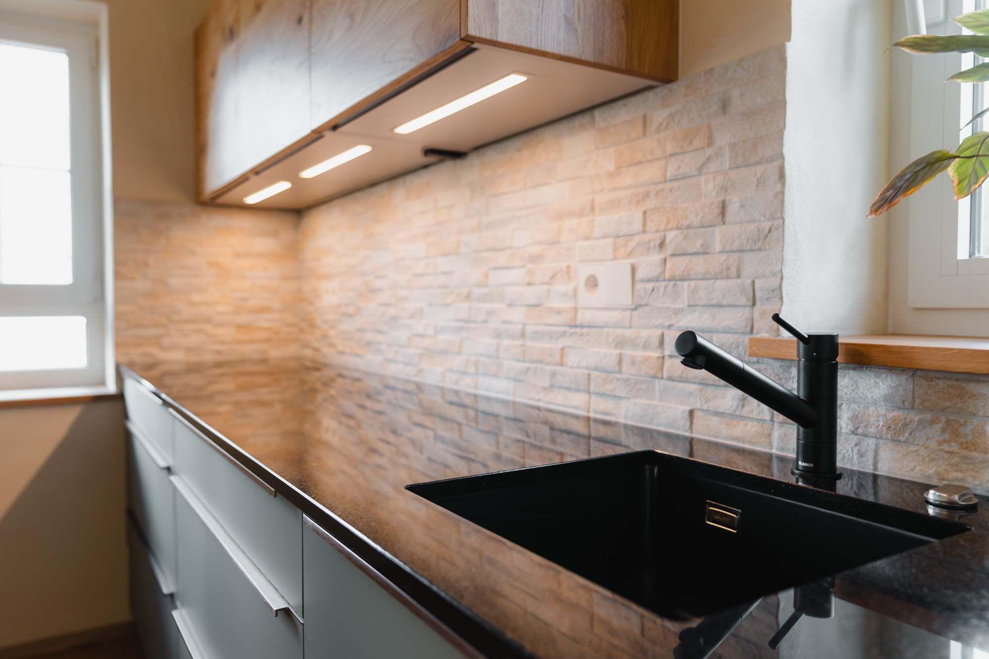 pülbereich mit schwarzer Unterbauspüle, schwarzer Armatur, Steinverblender-Rückwand und LED-Unterschrankbeleuchtung unter Holz-Oberschrank.