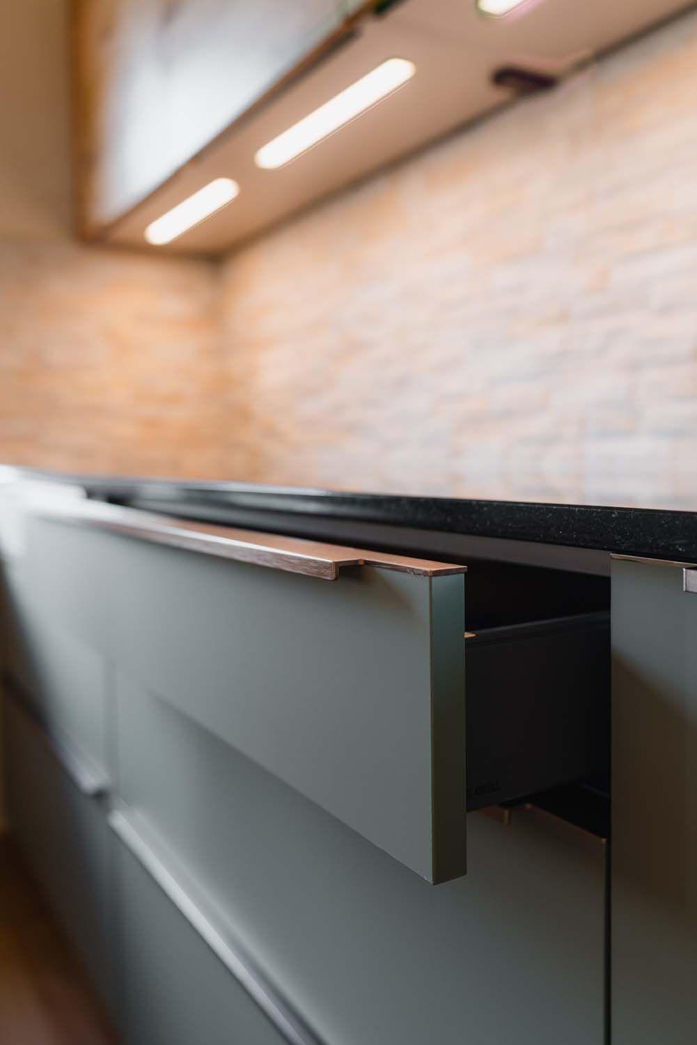 Geöffnete Schublade mit integriertem Holzgriff unter schwarzer Granitarbeitsplatte, unscharfe Steinverblender-Rückwand im Hintergrund.