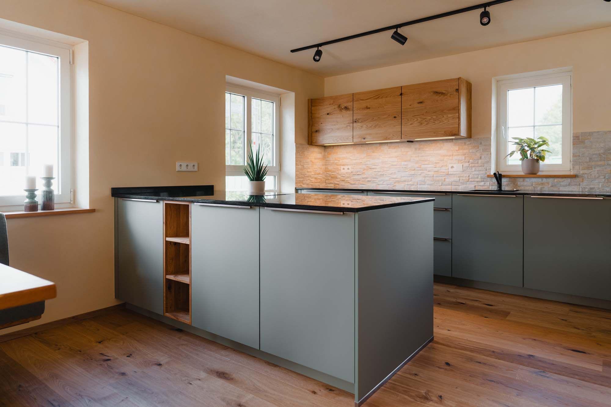 Freistehende Kochinsel mit offenem Holzregal-Element, salbeigrünen Fronten, schwarzer Arbeitsplatte, Holzoberschränke und Steinverblender-Rückwand im Hintergrund.