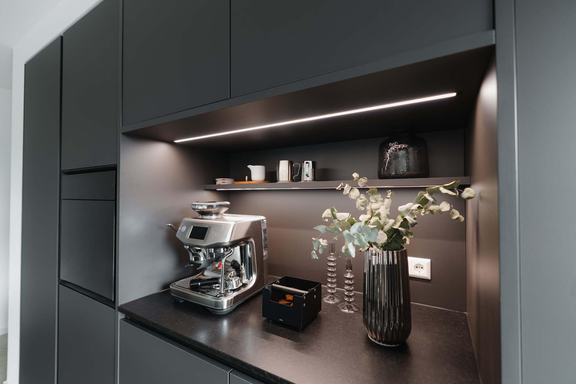 Beleuchtete Kaffeebar-Nische mit LED-Lichtleiste, Siebträgermaschine, Eukalyptus-Deko und schwarzer Rückwand unter grauen Oberschränken.