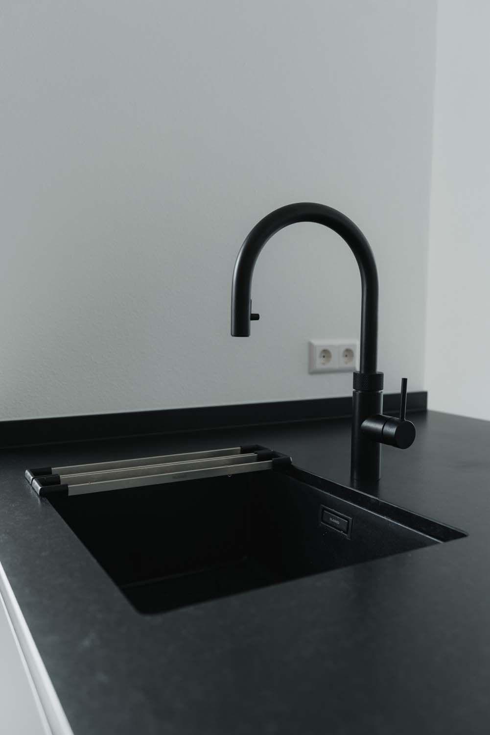 Nahaufnahme schwarze Unterbauspüle mit schwarzer Designer-Armatur, ausklappbarem Edelstahl-Abtropfgitter auf schwarzer Arbeitsplatte.