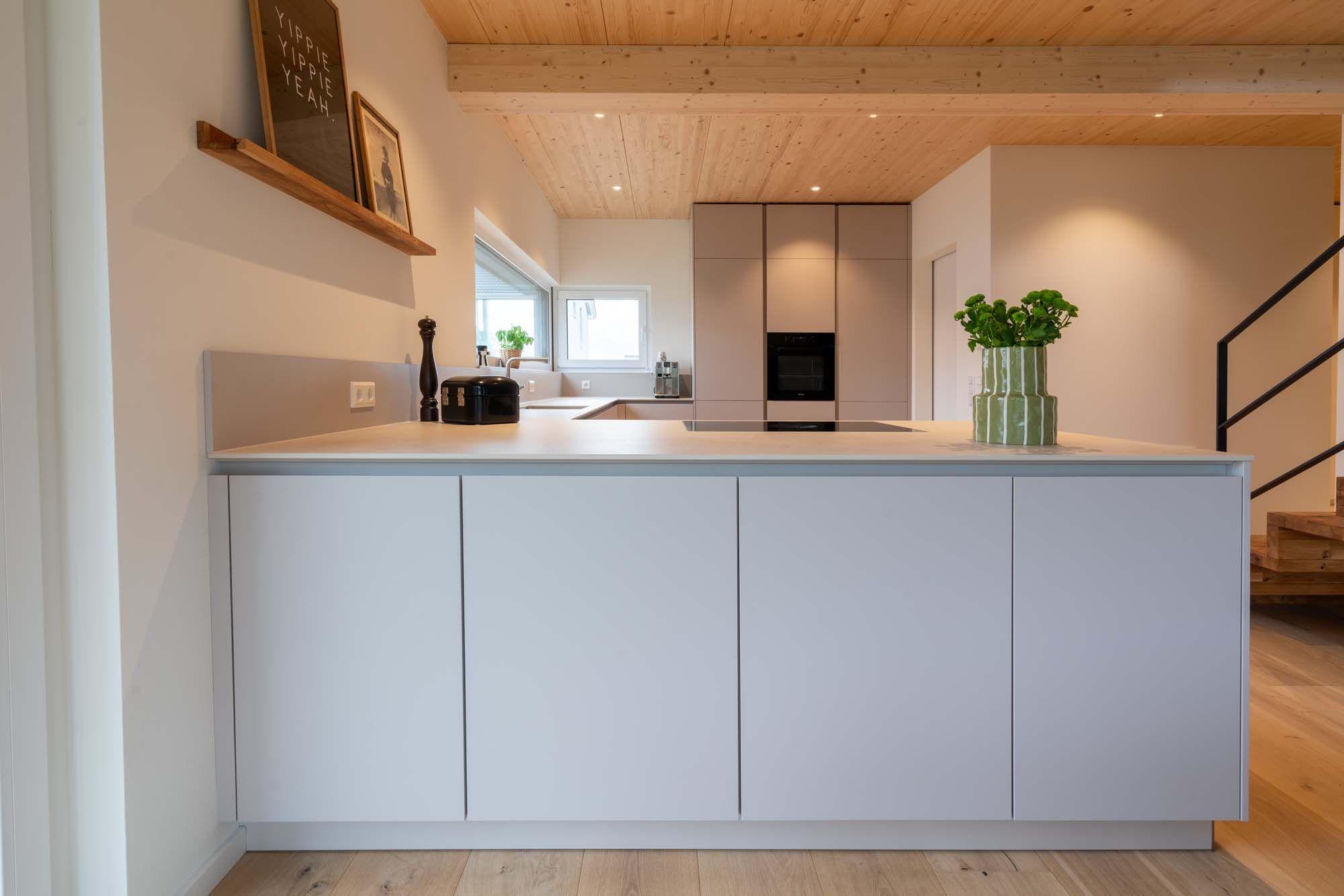 Kücheninsel mit minimalistischer Front vor beigefarbener Hochschrankwand mit Holzdecke.